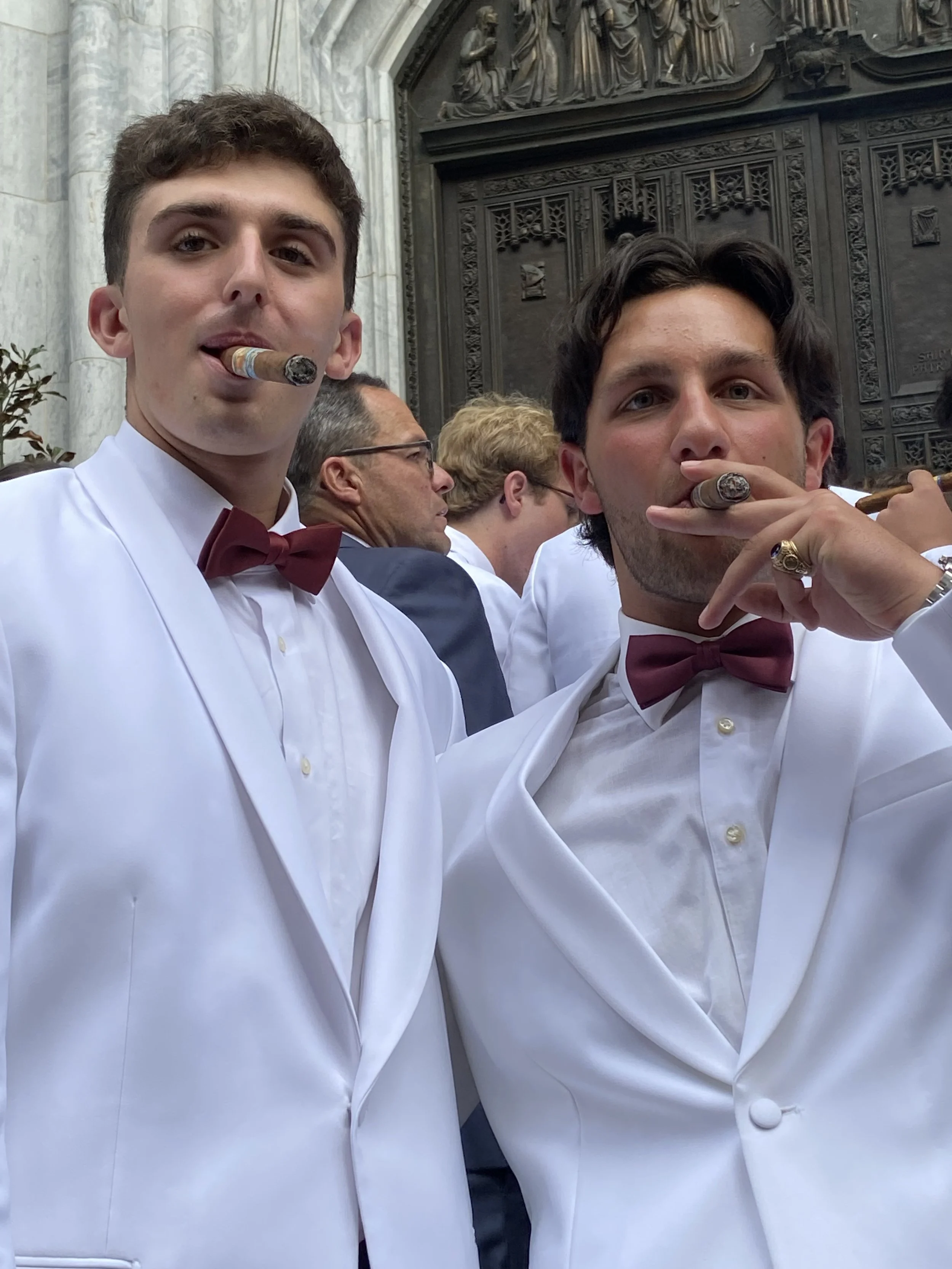 Two young men in white tuxedos with maroon bow ties, each smoking cigars, at a formal event with others in the background, standing in front of a large ornate door.