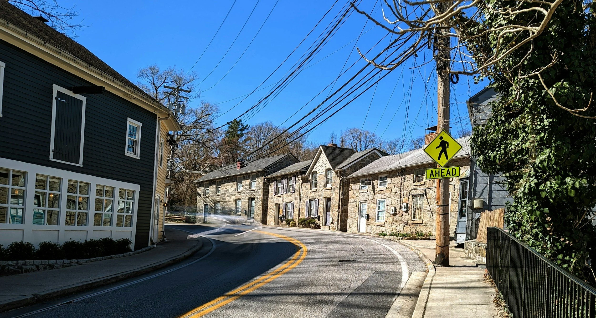 A curved street with houses on the right side and a sidewalk on the left. There is a yellow pedestrian crossing sign on a pole that says 'AHEAD' and shows a walking figure. Power lines run across the top of the image. The sky is clear and blue, with leafless trees in the background.