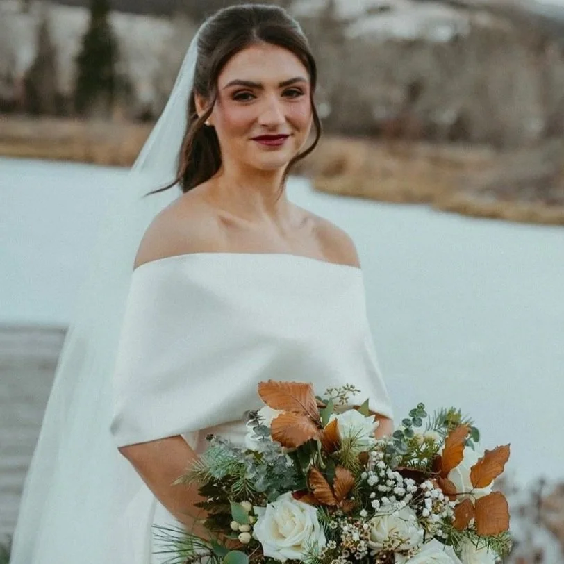 A woman dressed in an off-shoulder white wedding gown holding a bouquet with white roses and autumn leaves, outdoors near a water body. Millay and Meadowlark Flower Farm in Missoula, Montana. Weddings and Corporate events.
