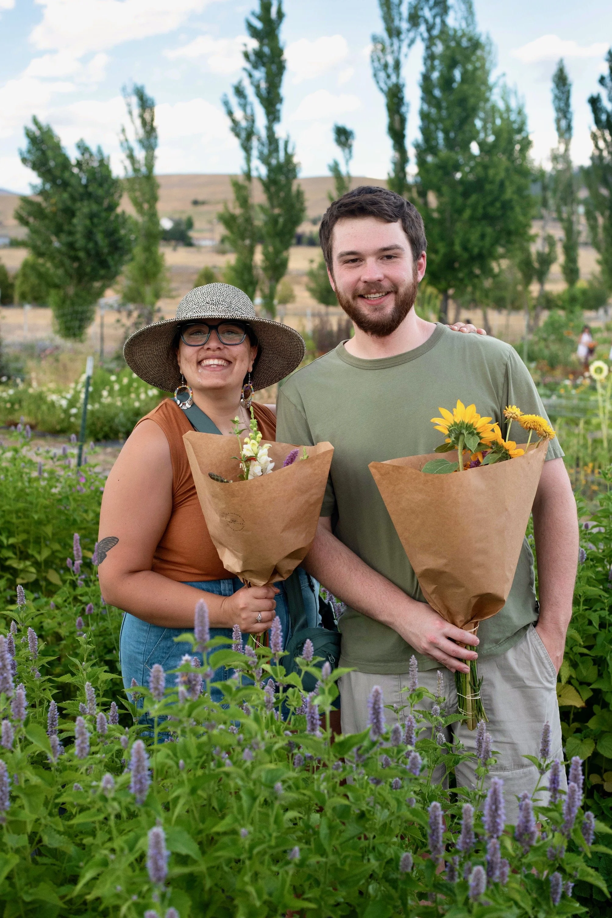 A smiling woman and man standing in a garden holding bouquets of flowers, with trees and distant hills in the background. Millay and Meadowlark Flower Farm in Missoula, Montana. Weddings and Corporate events.