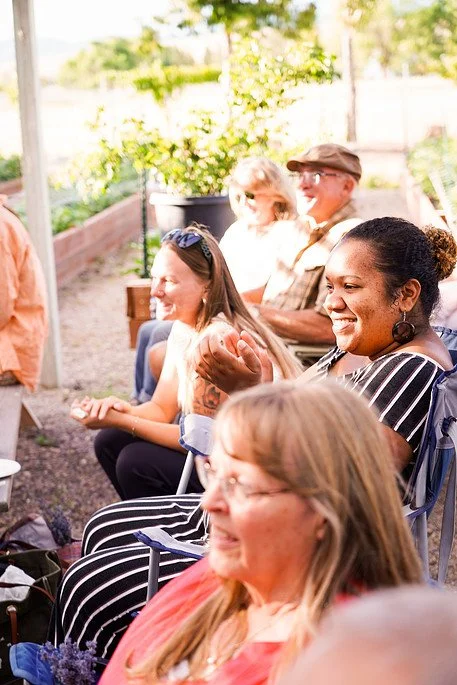 People sitting outdoors in a garden, smiling and clapping during a social gathering. Millay and Meadowlark Flower Farm in Missoula, Montana. Weddings and Corporate events.