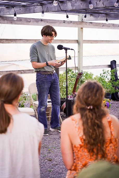 A young man reading from a book at a microphone in an outdoor setting, with an audience of women seated, some with long hair, and decorative string lights overhead. Millay and Meadowlark Flower Farm in Missoula, Montana. Weddings and Corporate events