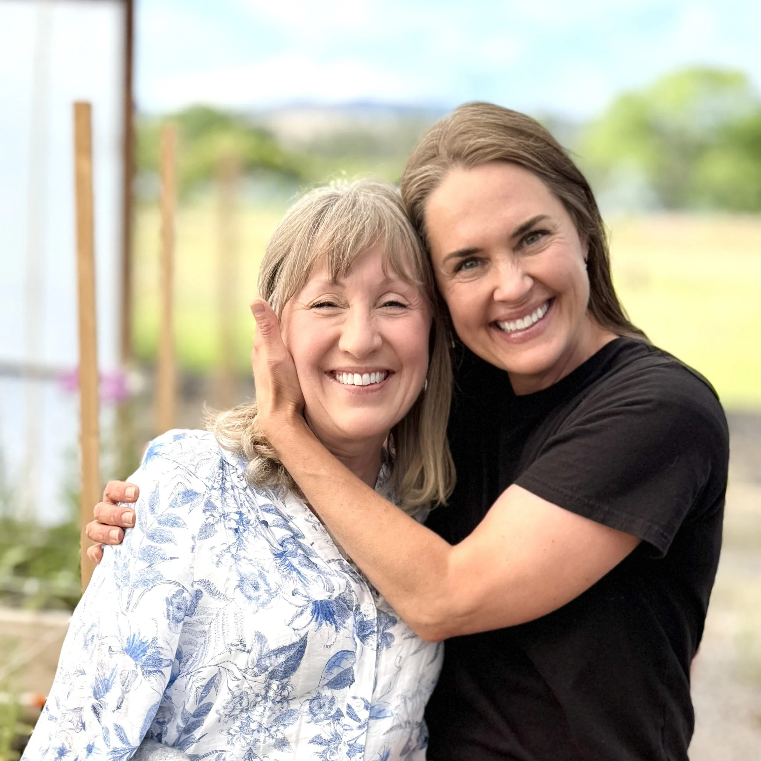Two women smiling and hugging outdoors, one with blonde hair in a floral blouse and the other with dark hair in a black t-shirt. Millay and Meadowlark Flower Farm in Missoula, Montana. Weddings and Corporate events.