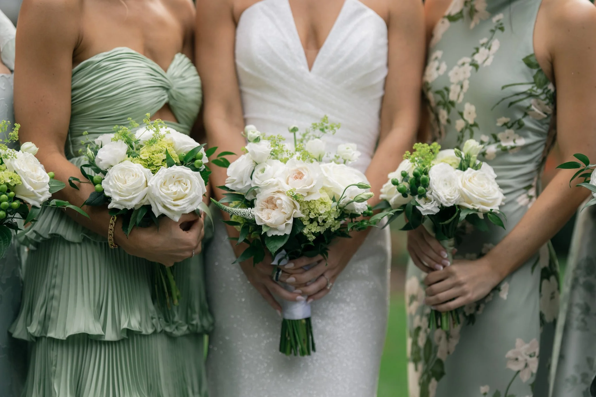Three women in wedding and bridesmaid dresses holding bouquets of white and pale pink flowers with greenery.