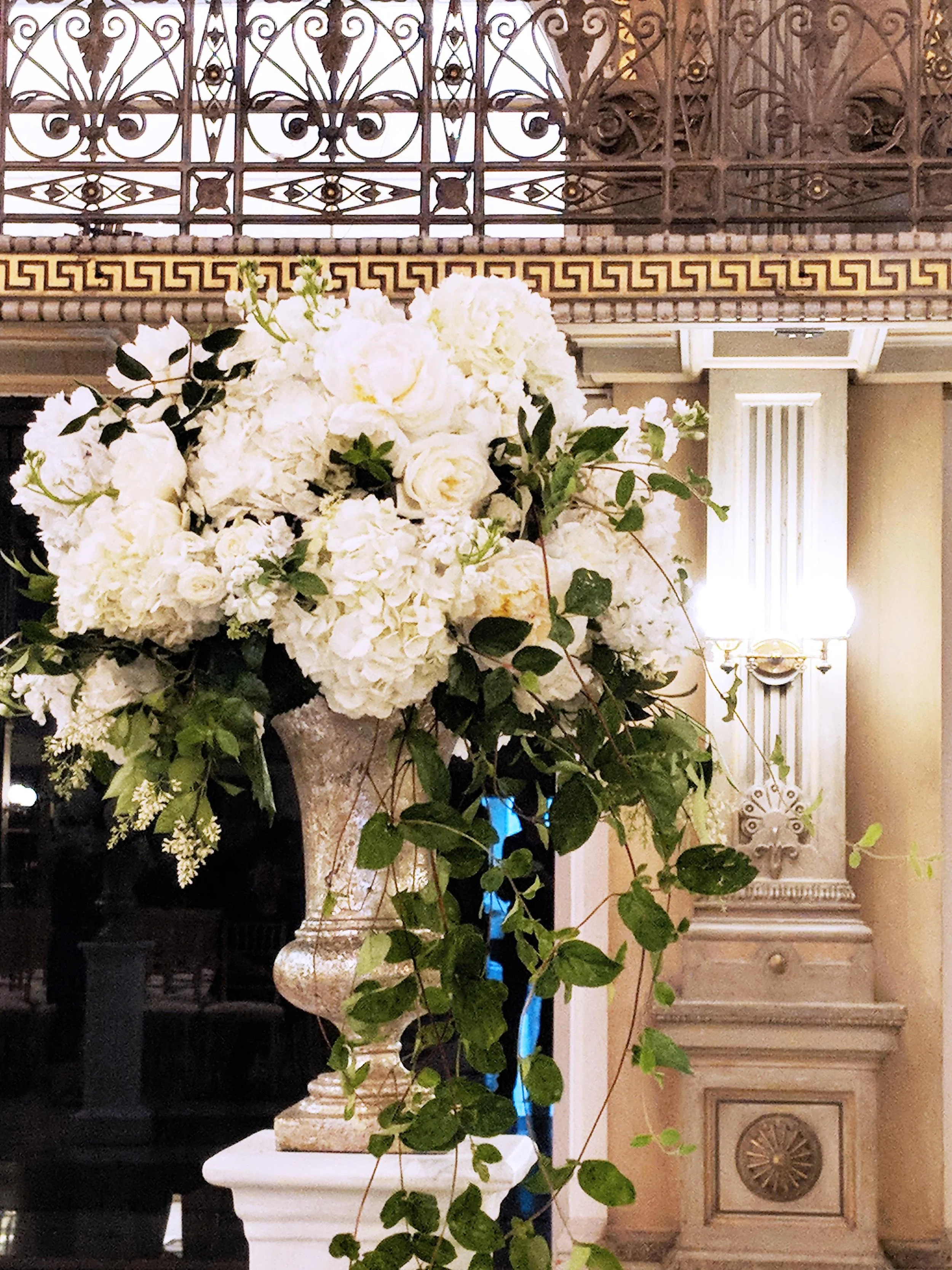 A large floral arrangement with white flowers, mostly roses and hydrangeas, with green leaves, in an ornate vase on a classical pedestal, inside a decorated elegant room with gold accents and a decorative railing.