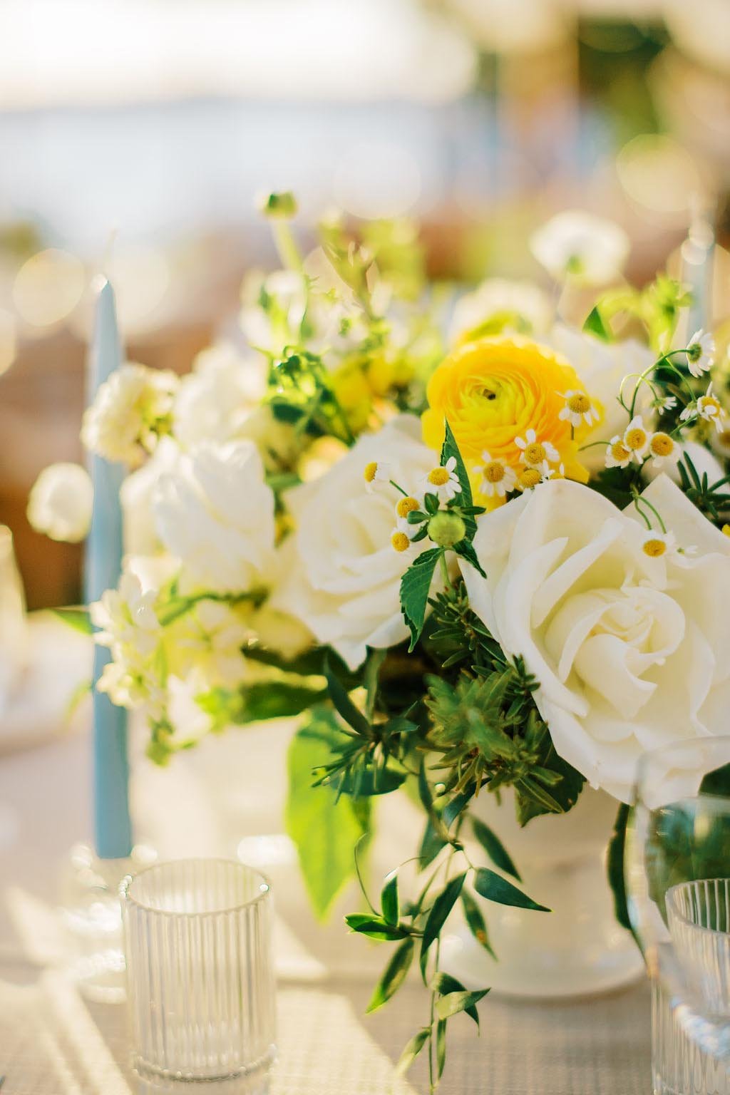 A floral centerpiece with white roses, yellow ranunculus, and small white daisies on a table, with blurred background and candle holder.