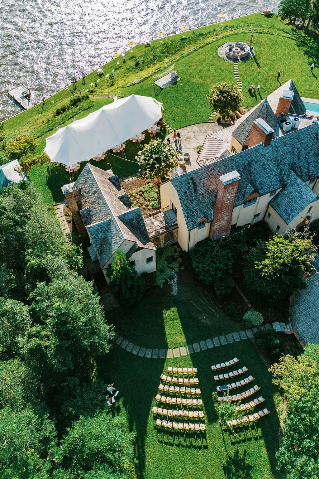 Aerial view of a house with a lush backyard setup for an outdoor event, featuring rows of chairs arranged on the grass, a white canopy tent, a fire pit area, and the waterfront in the background.