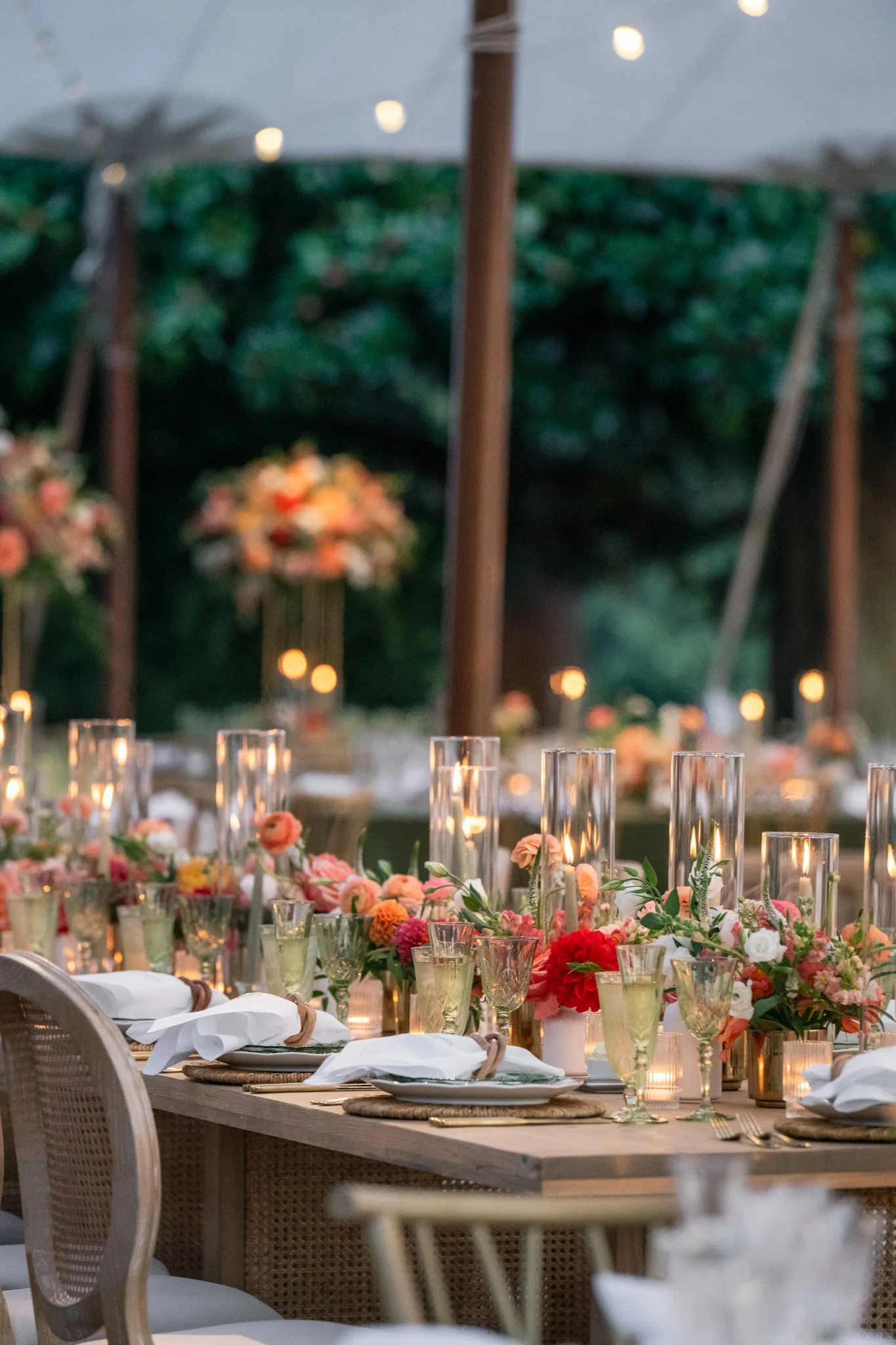 A beautifully decorated outdoor dining setup with flowers, candles, plates, glasses, and napkins under a tent.