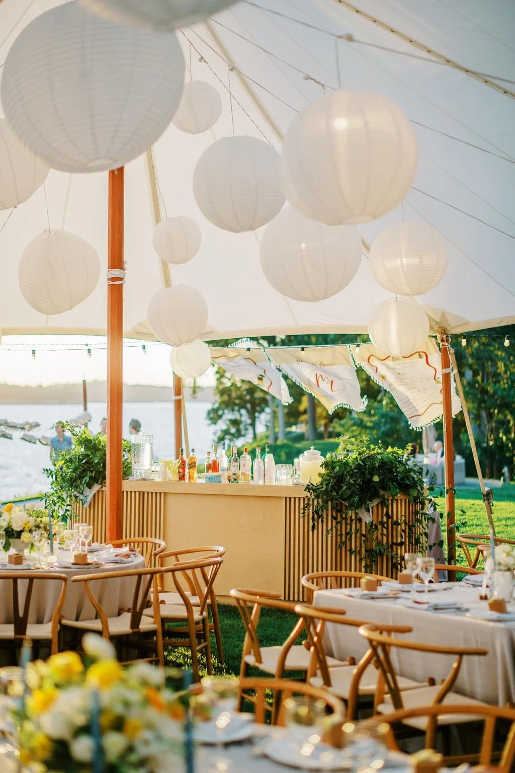 Outdoor event setup with white paper lanterns hanging from a tent, decorated tables with flowers and place settings, and a drink station with bottles and glassware, near a body of water.