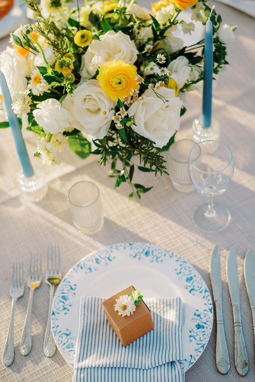 A decorated table with a floral centerpiece, blue candles, and a place setting featuring a striped napkin and small gift box.