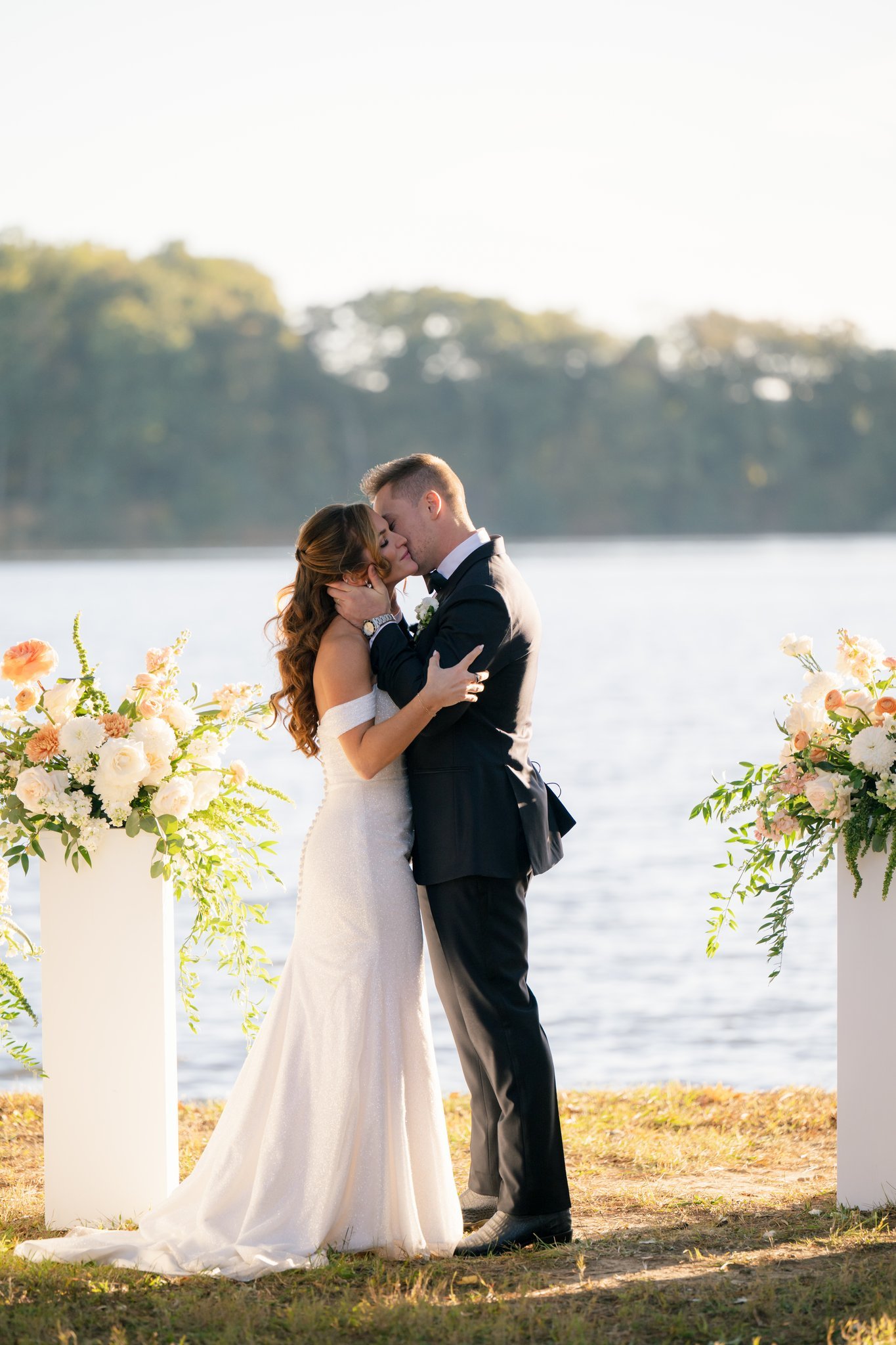 A bride and groom embracing during their wedding ceremony by a lake, with floral arrangements on tall white stands.
