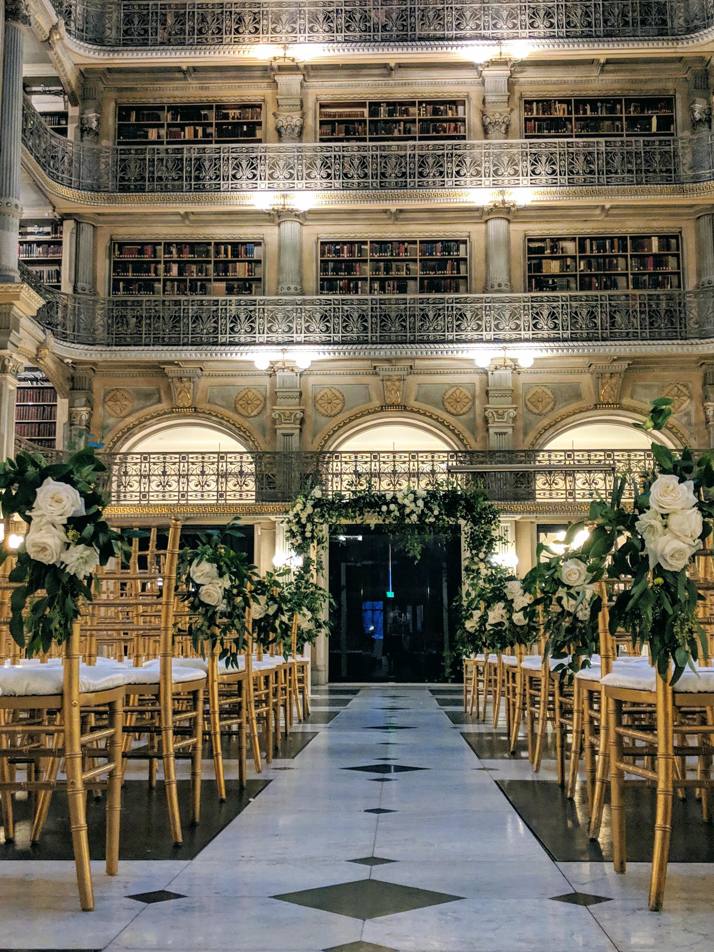 Elegant indoor wedding ceremony setup in a grand historic library or mansion with bookshelves, ornate railing balconies, floral arrangements, and gold Chiavari chairs.