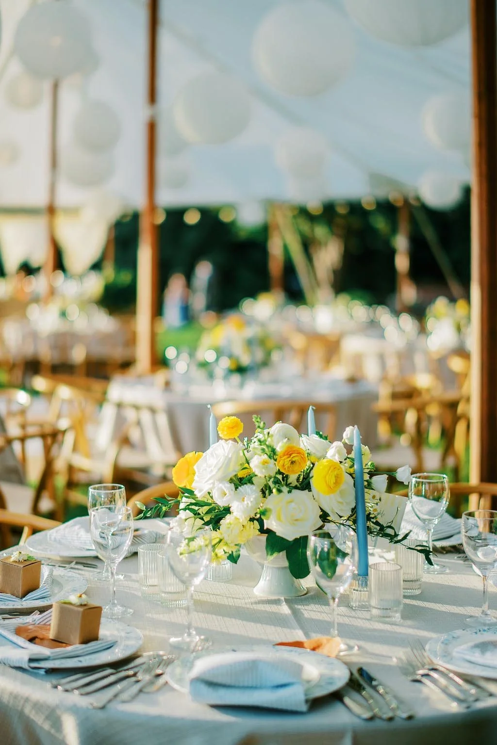 A table setting for an outdoor event with a floral centerpiece of white and yellow flowers, surrounded by wine glasses, plates, napkins, cutlery, and candles under a canopy with hanging paper lanterns.