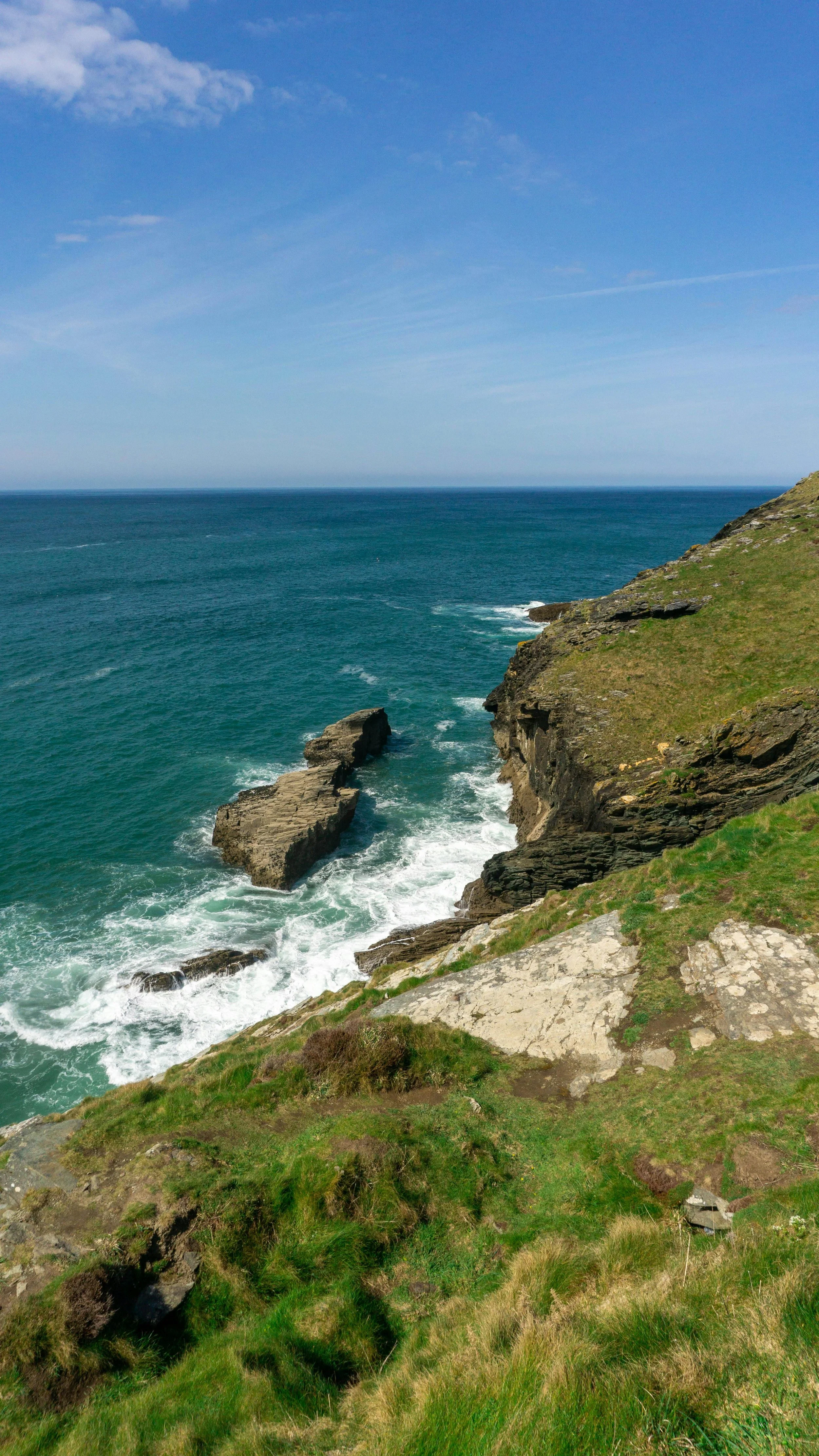 Cliffside view of the ocean with blue sky, clouds, and green grassy slope, rocky shoreline, and waves crashing against rocks.