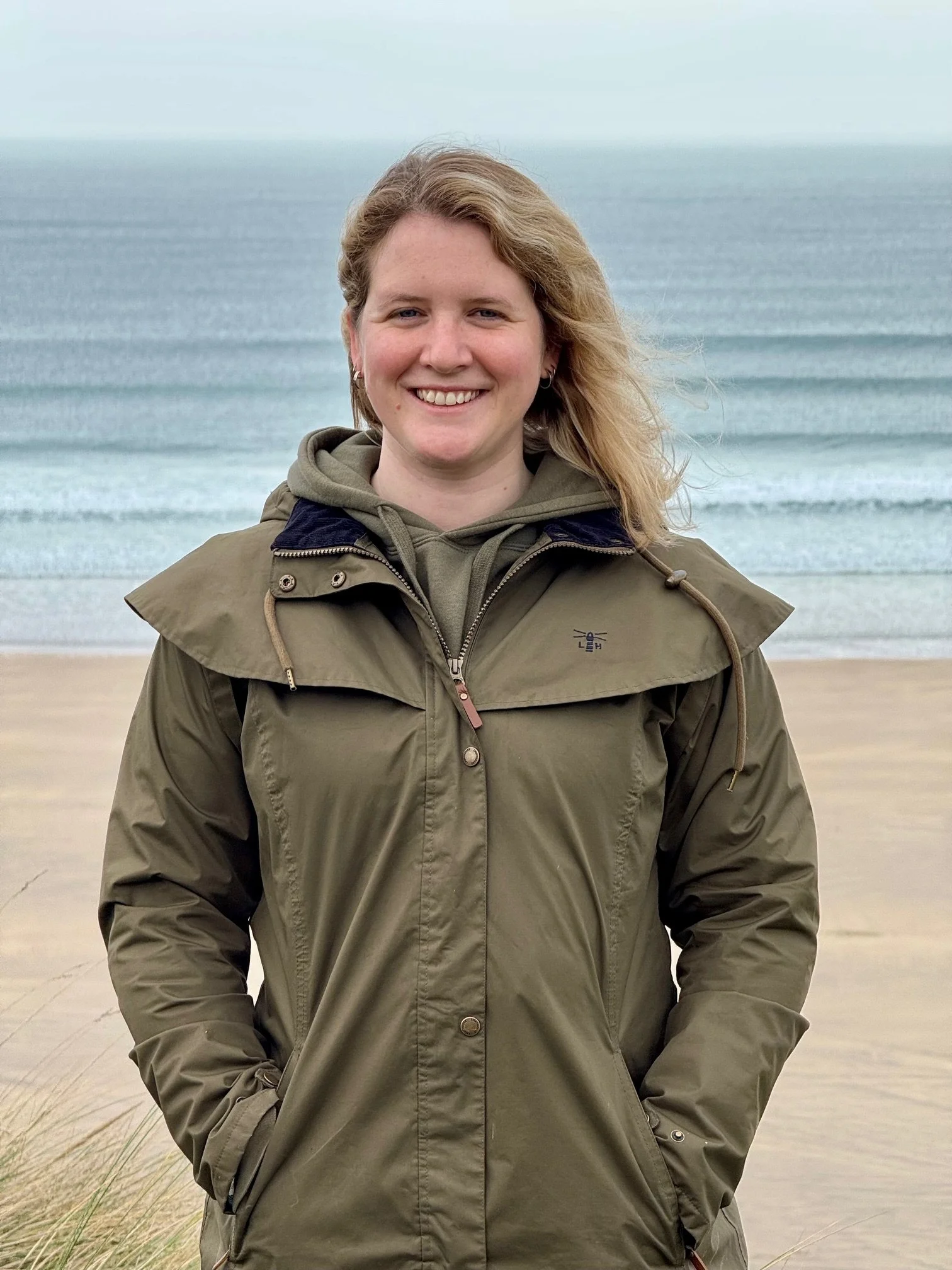 Abi, therapist and counsellor in West Cornwall, smiling at the camera outdoors on a beach, wearing a green jacket with a natural background of sand, grasses, and ocean waves.