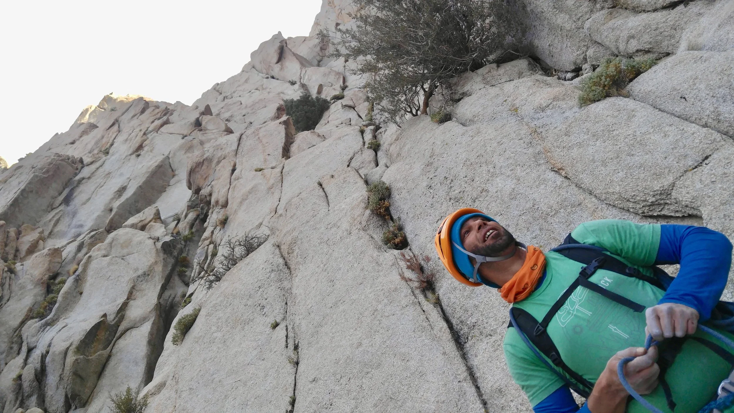 A man in climbing gear, including a helmet and harness, smiling while rock climbing on a steep granite cliff.