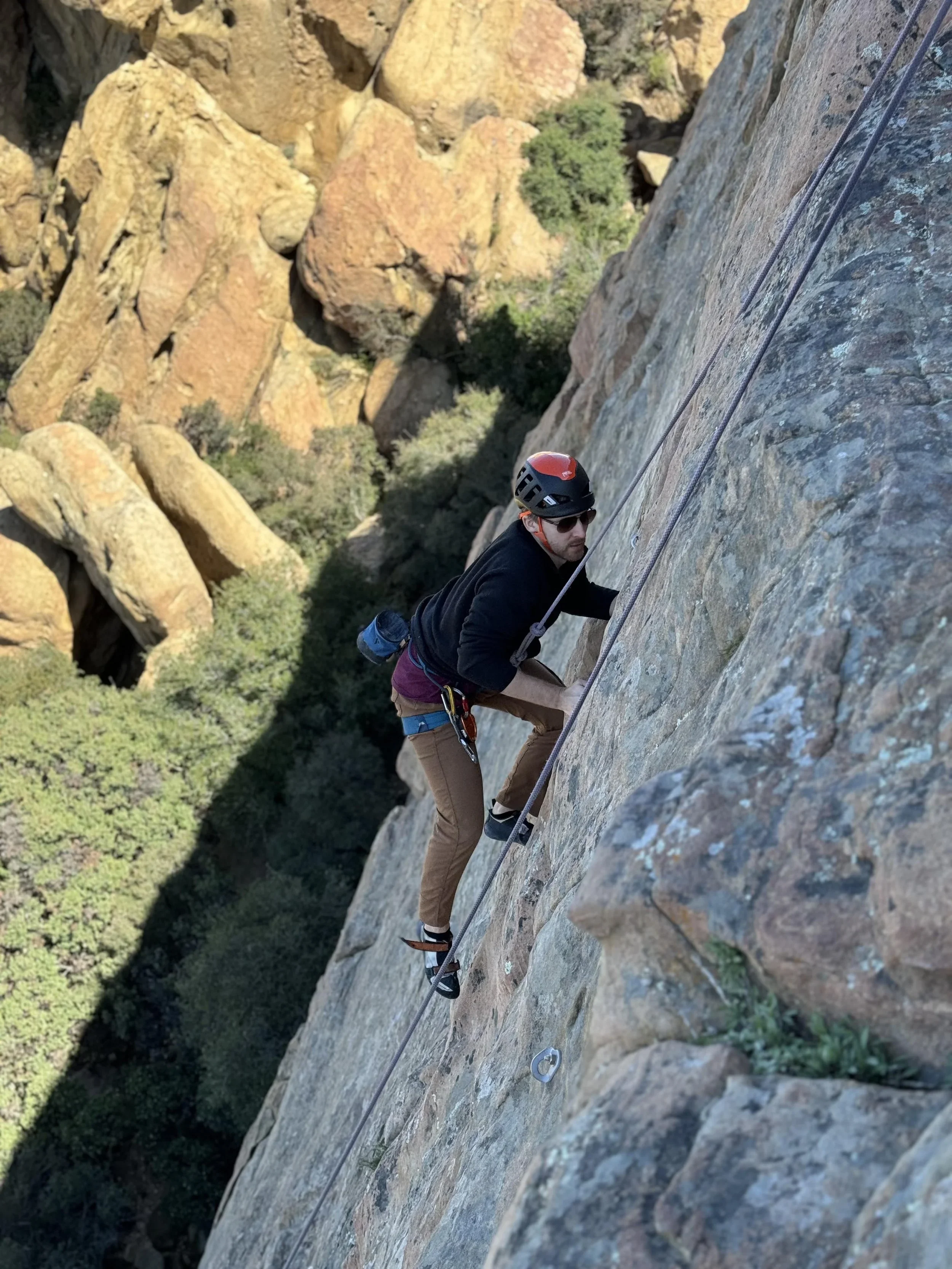 A person rock climbing on a steep, rocky cliff face with safety gear, including a helmet, harness, and ropes.