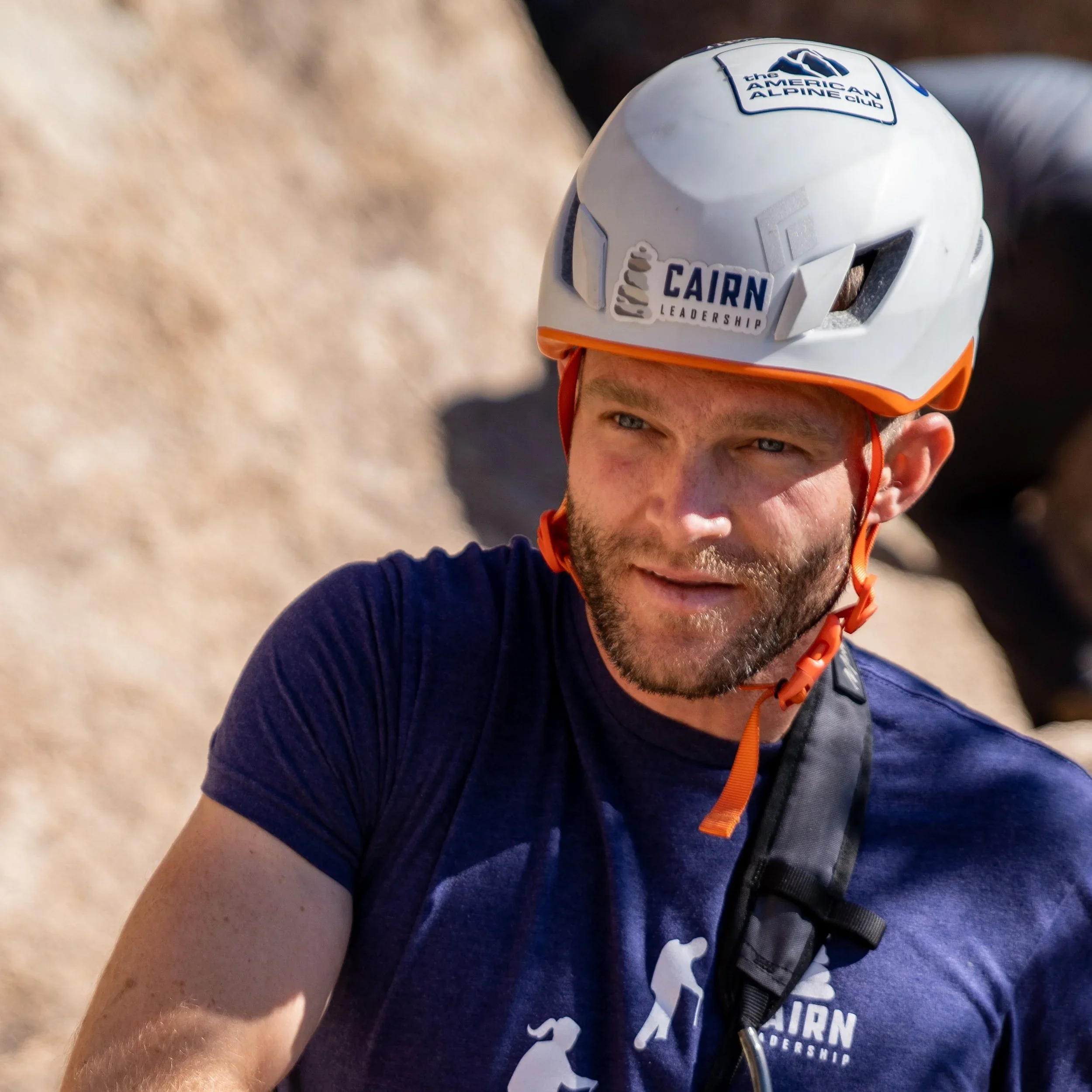 A man wearing a white helmet with outdoor adventure logos, including 'the American Alpine Club' and 'CAIRN Leadership,' is seen outdoors against a rocky background. He has a beard and is wearing a dark blue t-shirt with a climbing or outdoor logo.