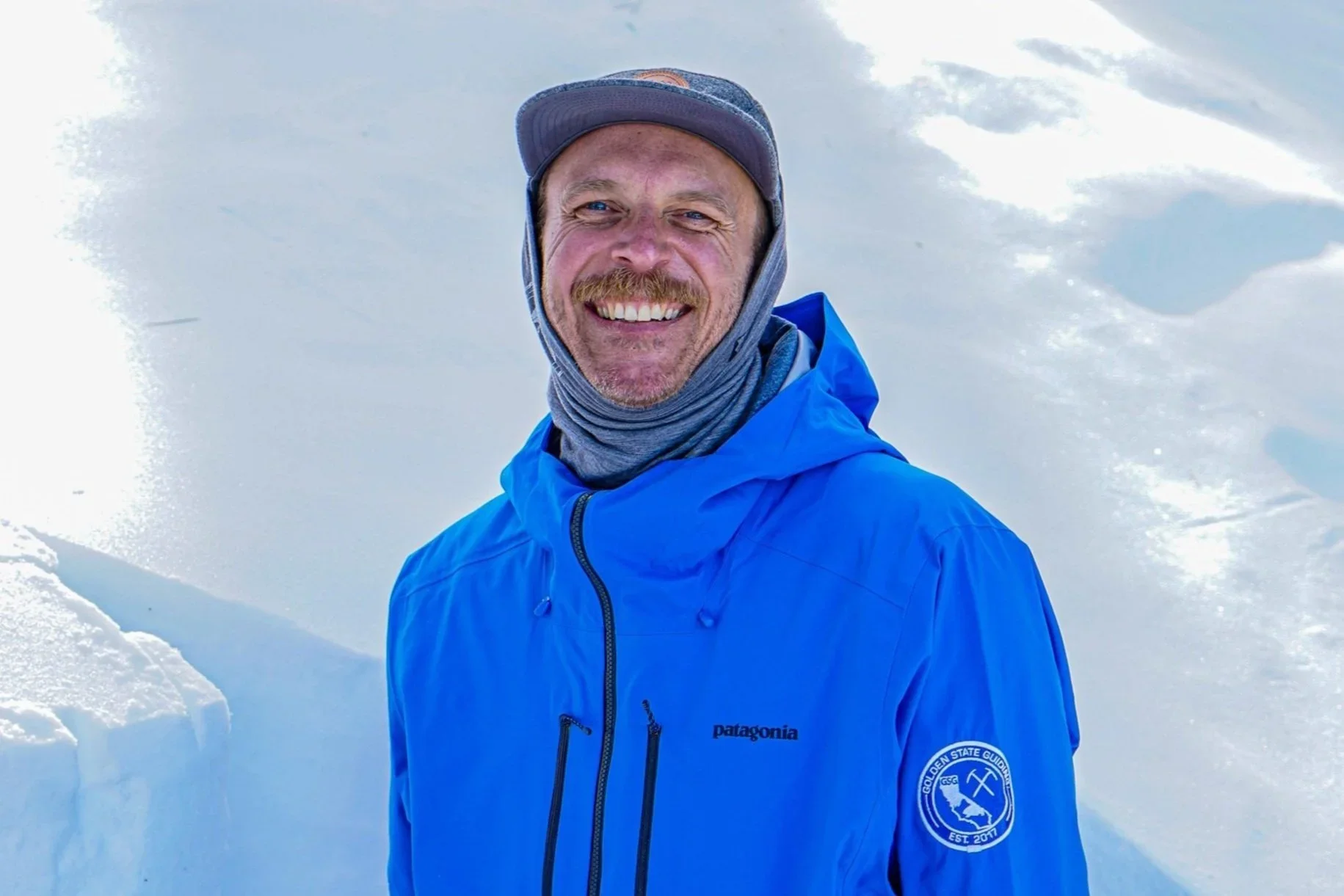 A man smiling outdoors in snowy weather, wearing a blue Patagonia jacket and a gray hat.