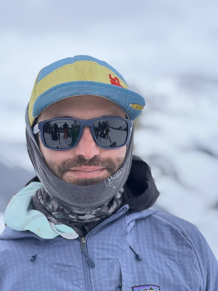 Man wearing a blue jacket, a yellow and blue cap, sunglasses, and a neck gaiter in a snowy outdoor setting.