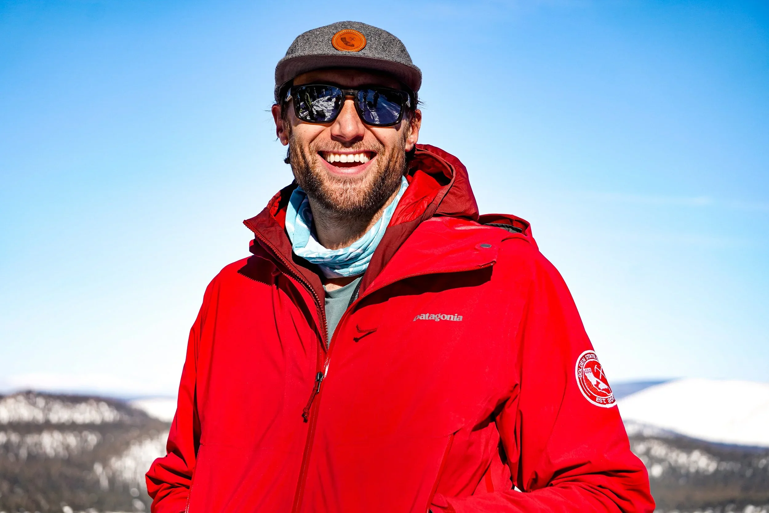 Man smiling wearing a red Patagonia jacket, sunglasses, a gray cap, and a neck gaiter outdoors with snow-covered mountains in the background.