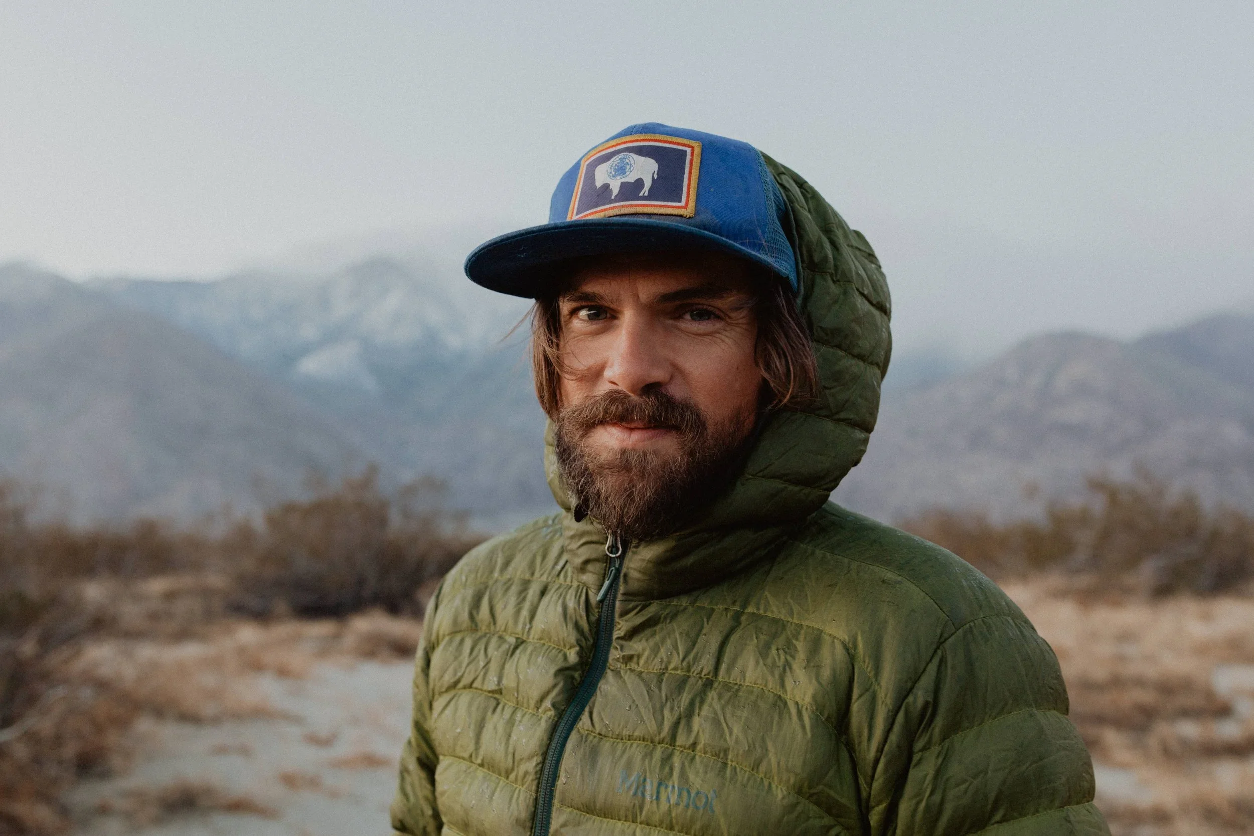 A man with a beard wearing a green puffer jacket and a blue cap with an animal patch, outdoors with mountains in the background.