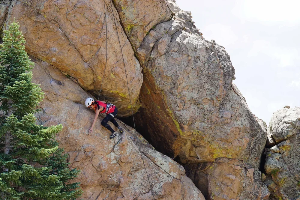 A person wearing a helmet and harness rock climbing on a large, rugged rock formation with a tree nearby.