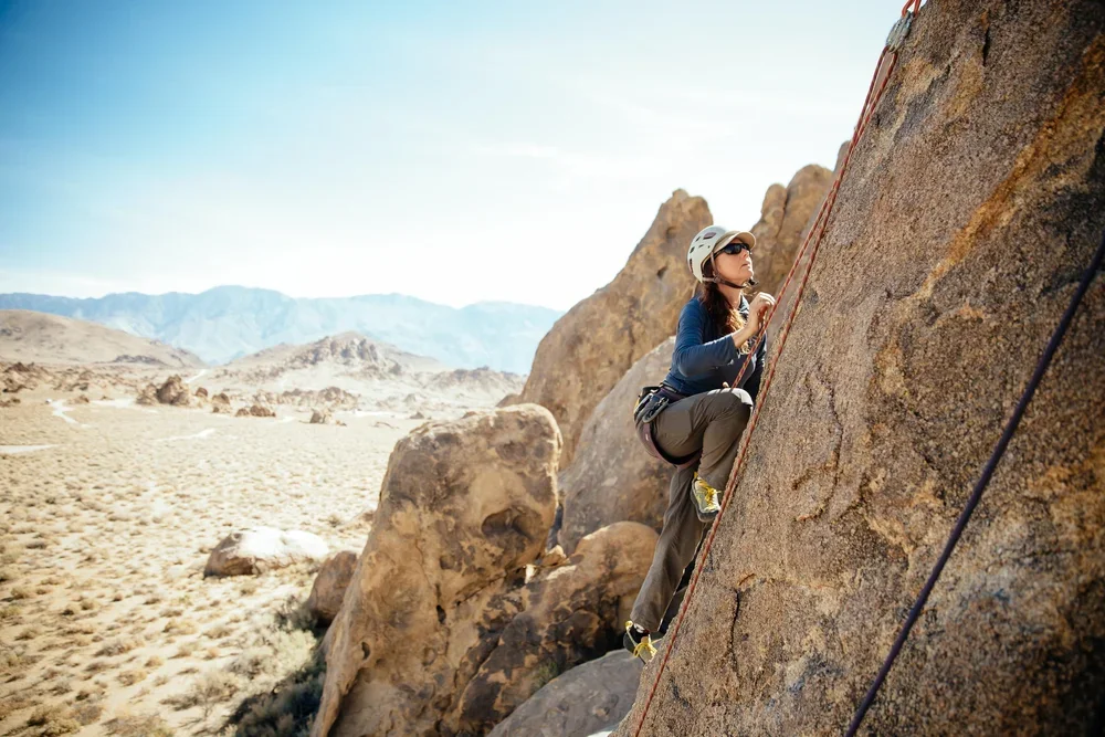 A woman rock climbing outdoors in a desert landscape, wearing a helmet and sunglasses.