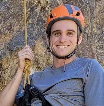 Man rock climbing outdoors, holding a yellow safety rope, smiling, wearing a helmet and a blue shirt.