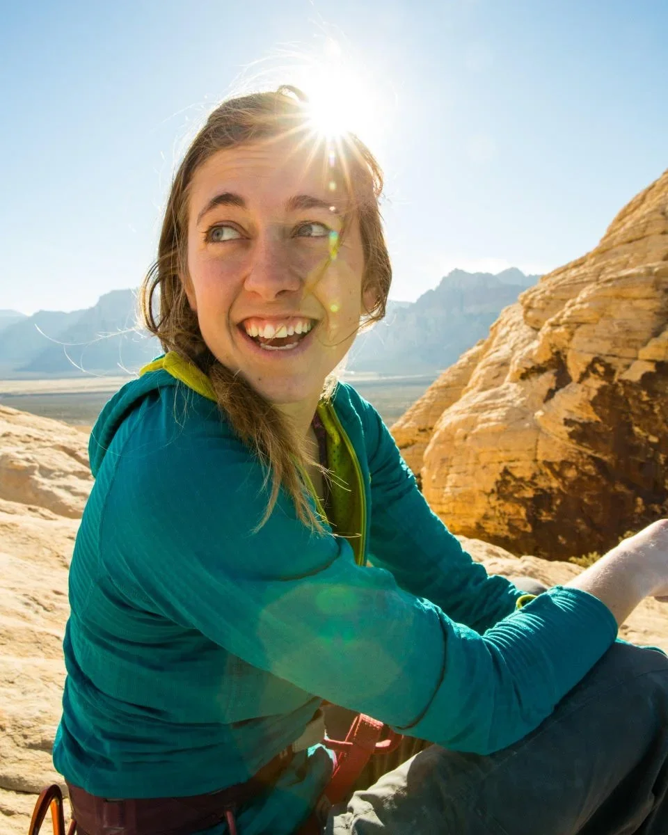 A woman with light brown hair smiling outdoors in bright sunlight, with rocky desert landscape and mountains in the background.