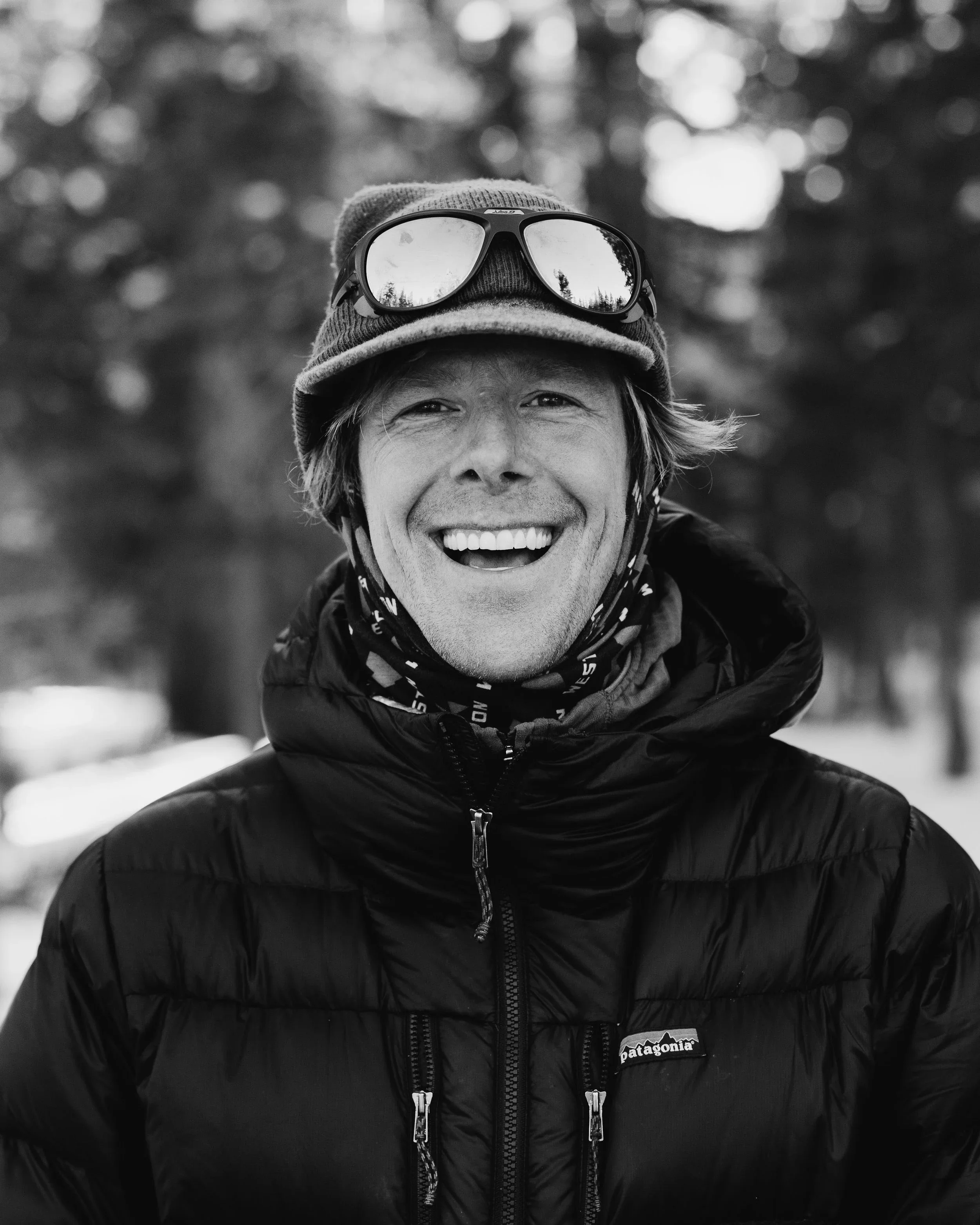 A man smiling, wearing a beanie hat, sunglasses on top, a scarf, and a puffy jacket outdoors in a park with trees in the background.