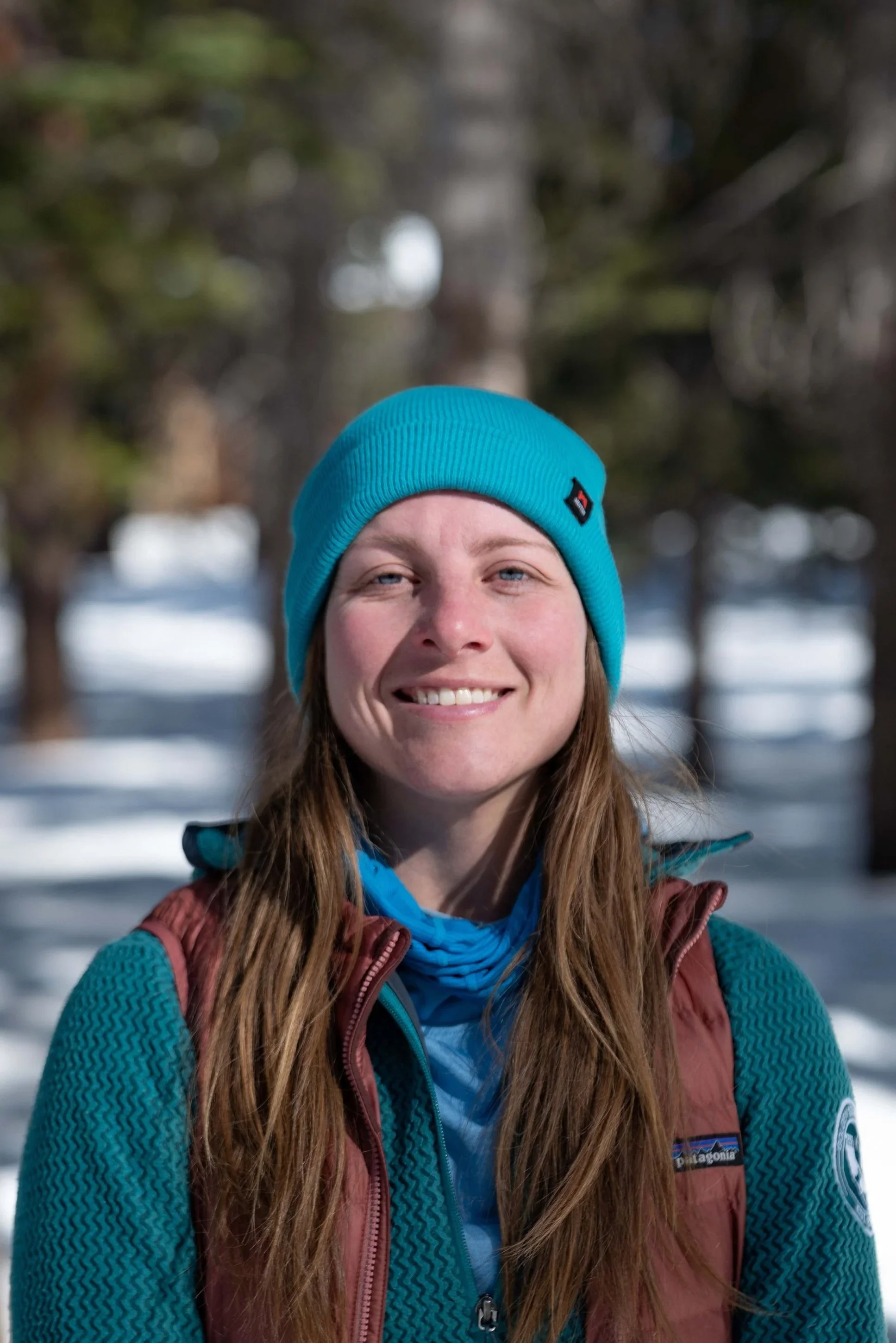 A woman outdoors in winter, wearing a blue beanie hat and a Patagonia jacket, smiling with trees and snow in the background.