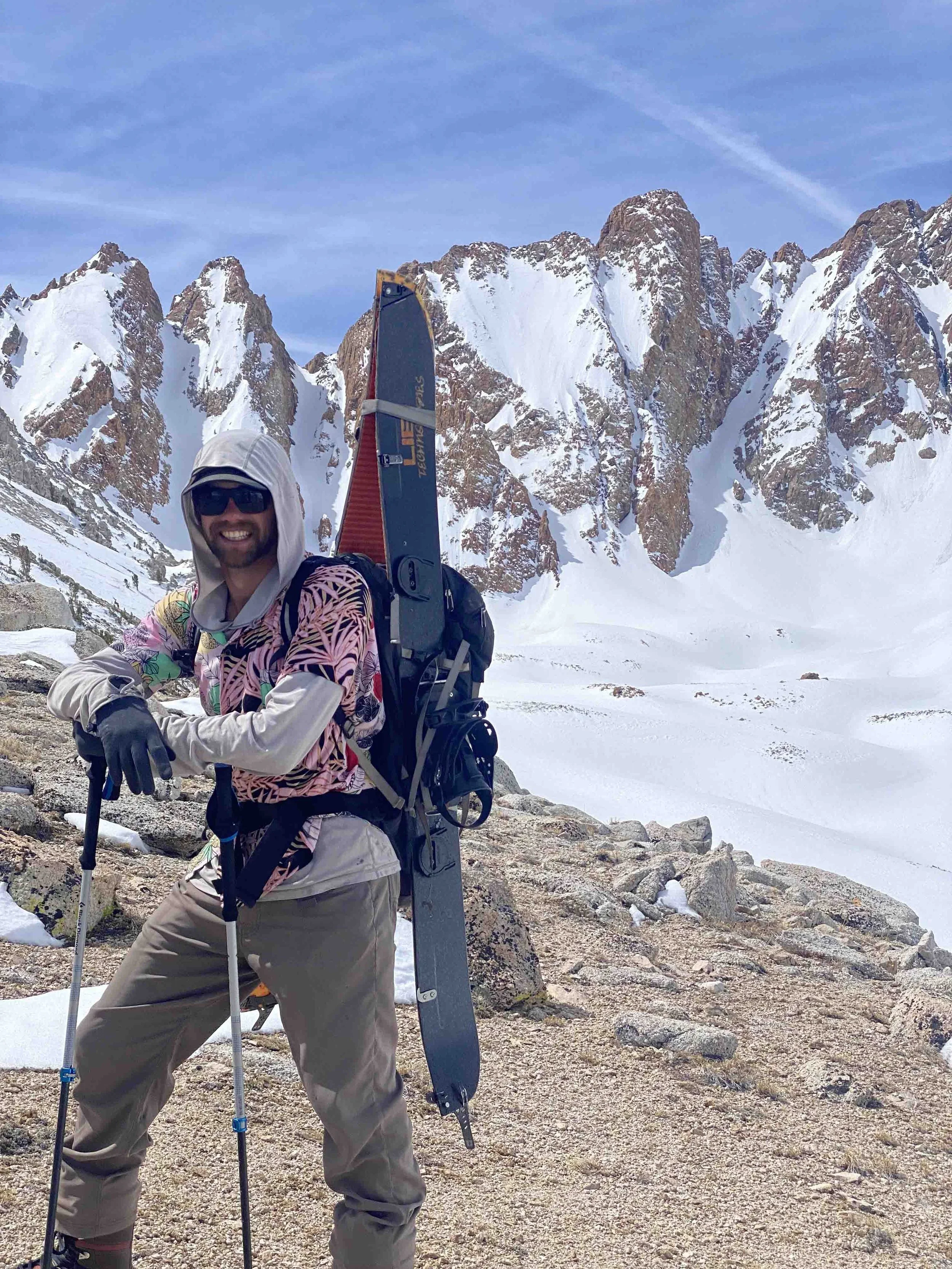 A man dressed in outdoor gear, smiling, standing on a rocky snow-covered mountain trail with snow-capped peaks in the background.