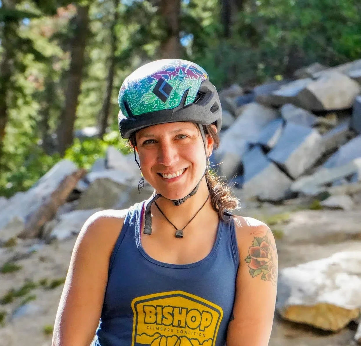 Smiling woman wearing a helmet and tank top with a camp logo, standing outdoors with trees and rocks in the background.