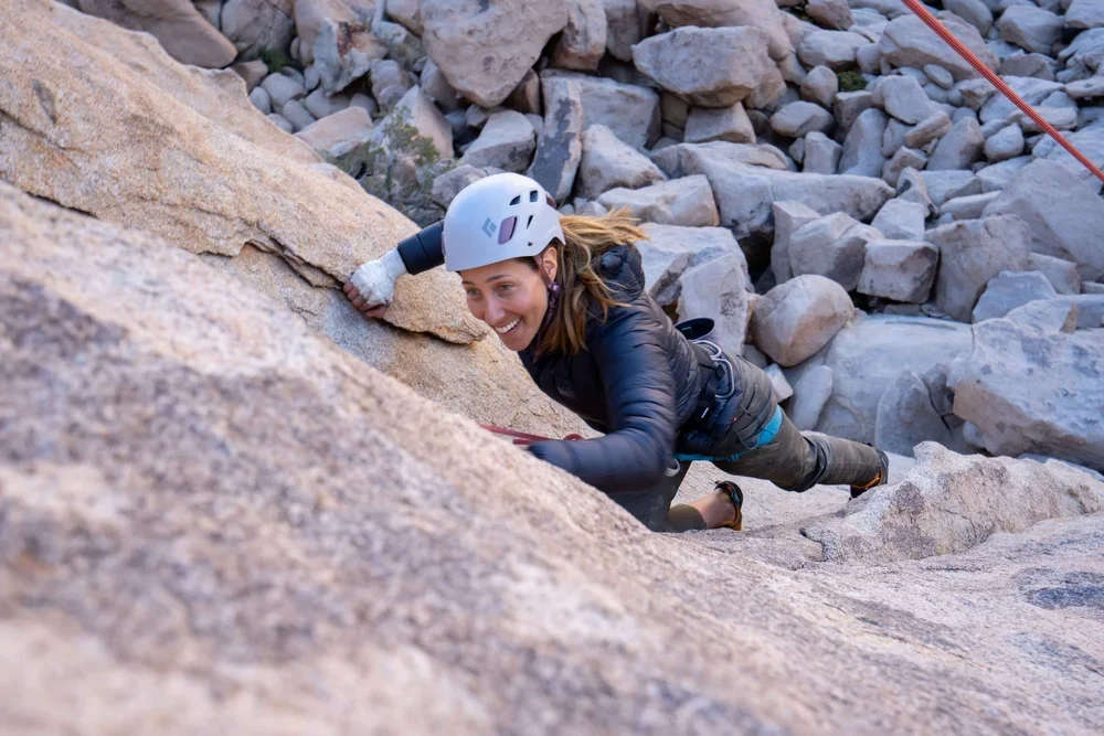 A woman in a helmet and outdoor gear climbing a steep rock face while smiling.