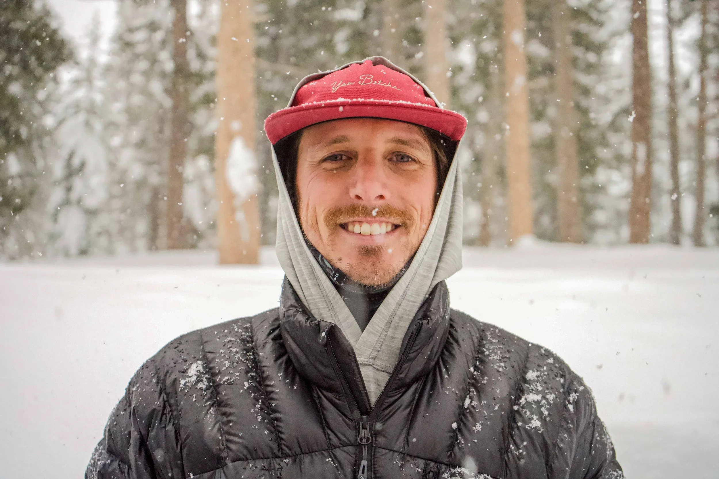 A man with light skin, a beard, and a mustache smiling outdoors in a snowy forest, wearing a red cap, a hooded sweatshirt under a black puffer jacket, with snow on his clothing and face.