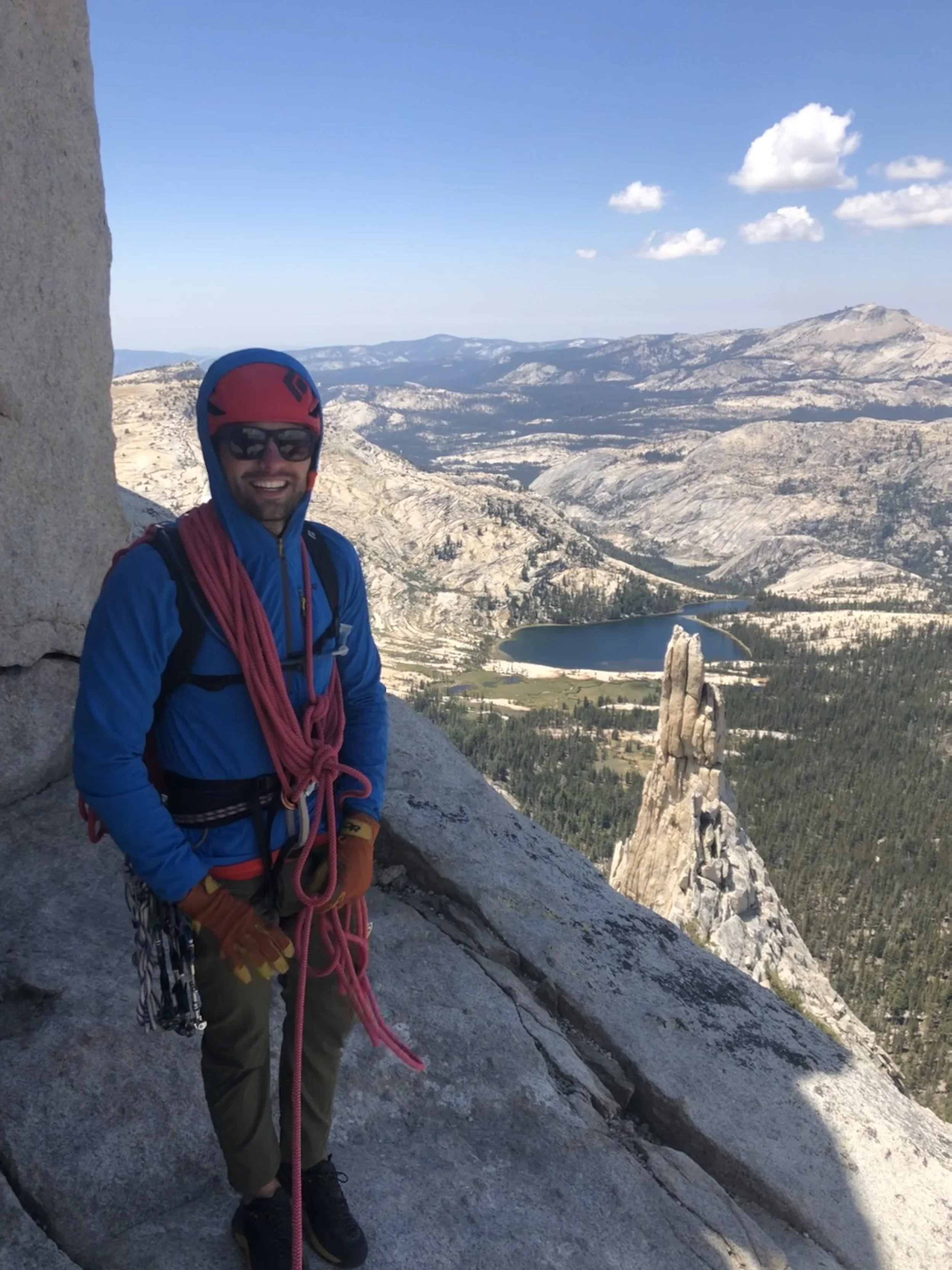 A man in climbing gear, including a helmet, harness, and gloves, standing on a rock ledge on a mountain, with a scenic view of rugged mountains, a lake, and a rock formation in the background.