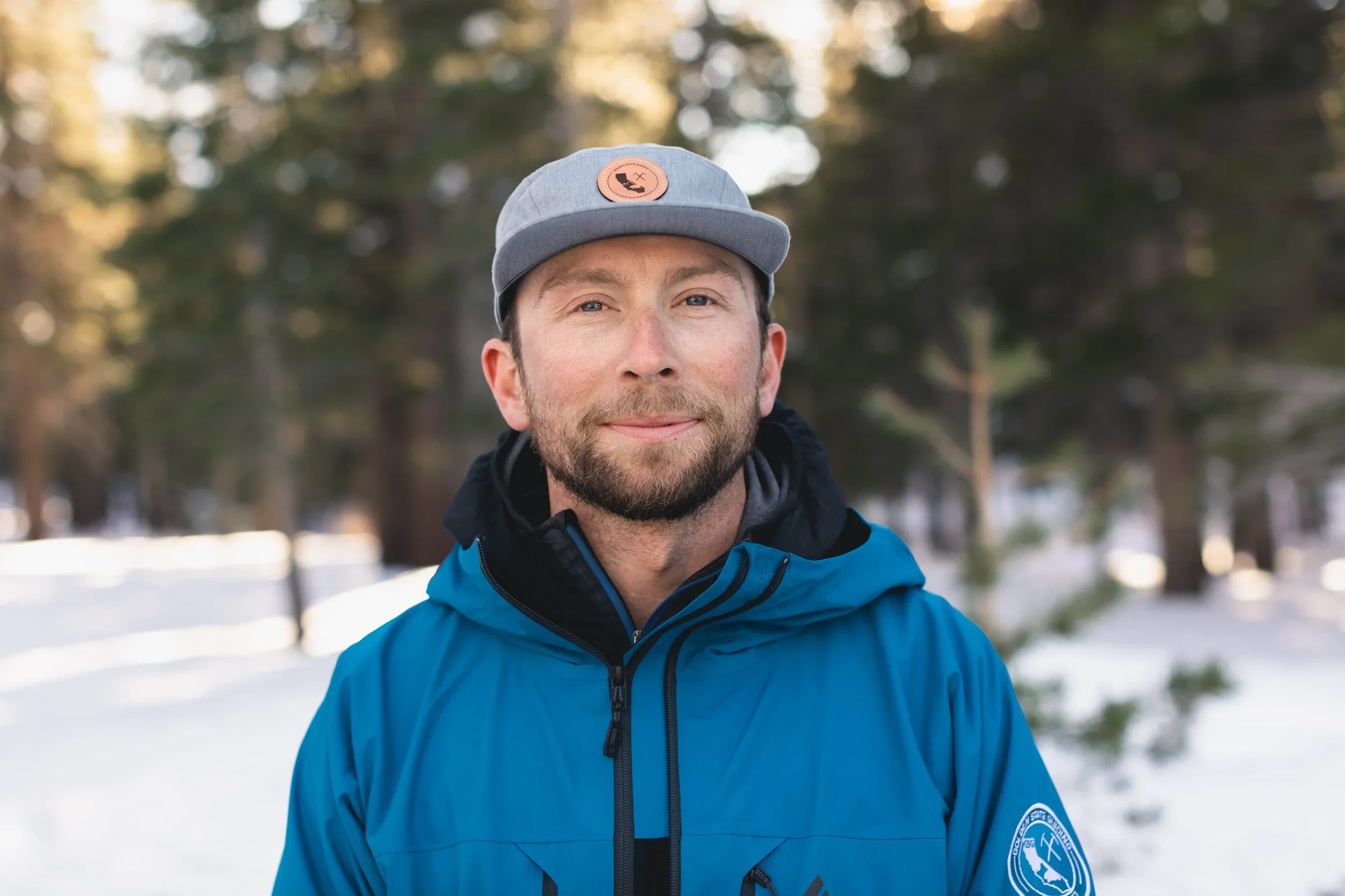 A man outdoors in a snowy forest, wearing a gray cap with a patch, a blue jacket, and a black hoodie.