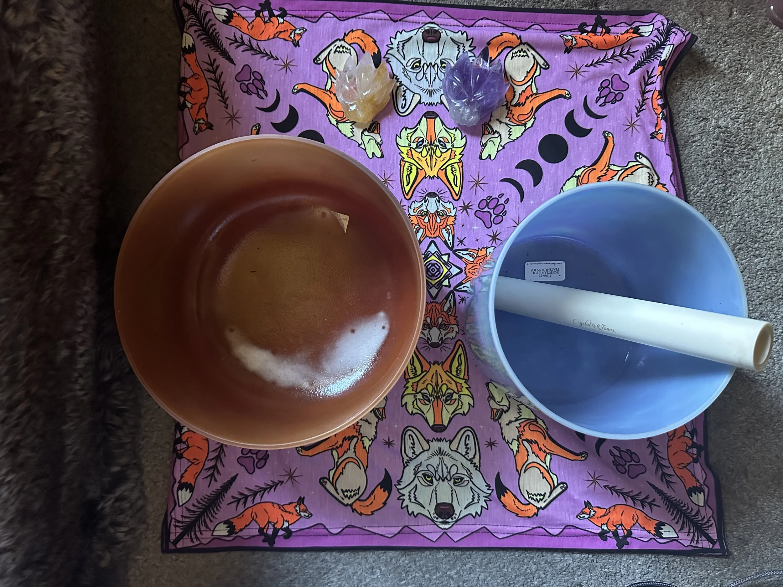 Coloring or meditation setup with a purple cloth featuring wolf, fox, and other animal illustrations, two semi-precious stone carvings, a large terracotta bowl, a large blue bowl with a white tube inside, positioned on a carpeted floor.