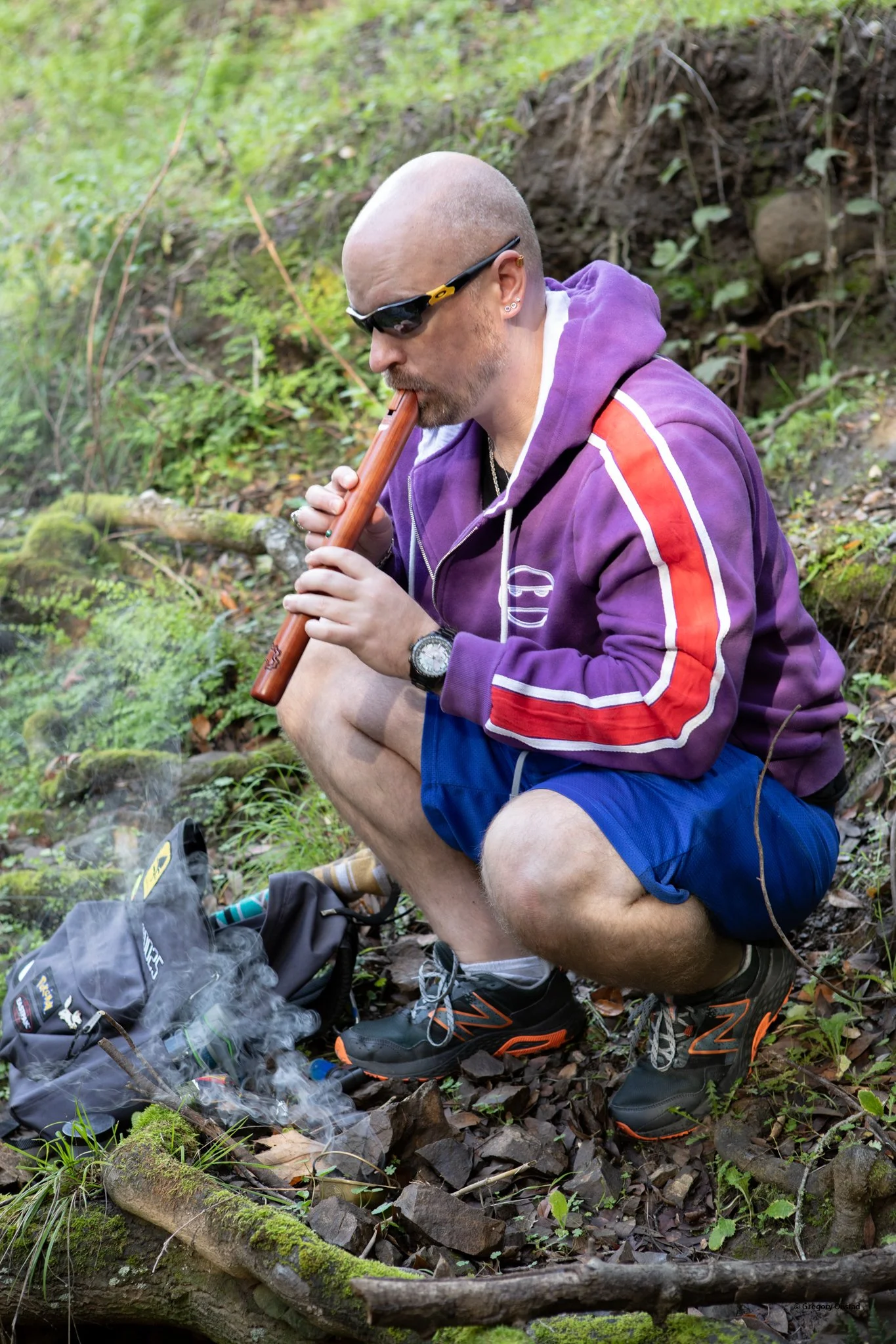 A man crouching outdoors in a wooded area, playing a wooden flute with smoke rising nearby, wearing a purple hoodie with red and white stripes, blue shorts, sunglasses, and athletic shoes.