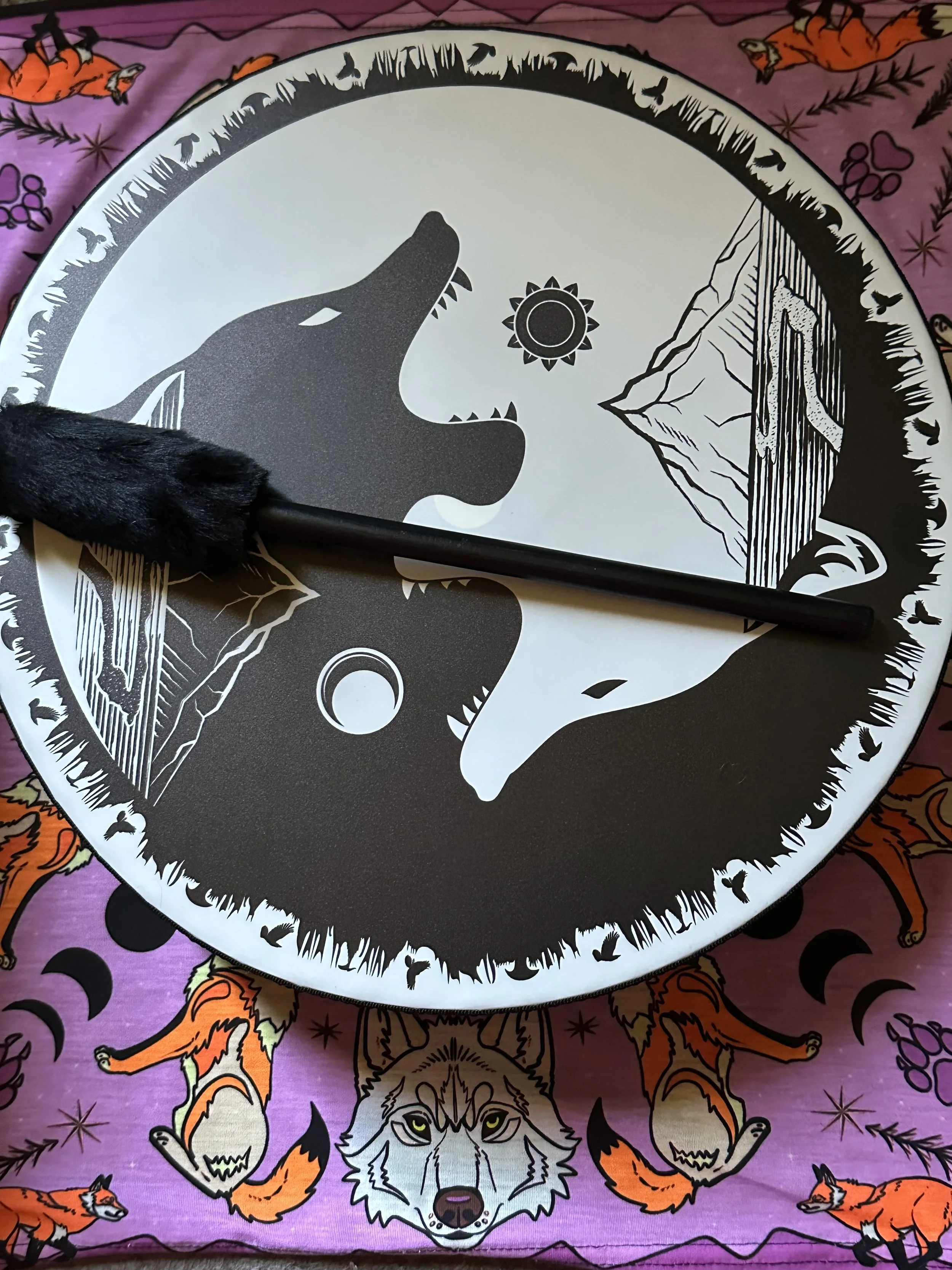 A black and white illustration of two wolf heads facing each other on a circular surface, with mountains and a sun in the background. There is a black stick with a furry end placed on the surface. The surface is on a colorful, patterned fabric featuring foxes, a wolf, and moon phases.