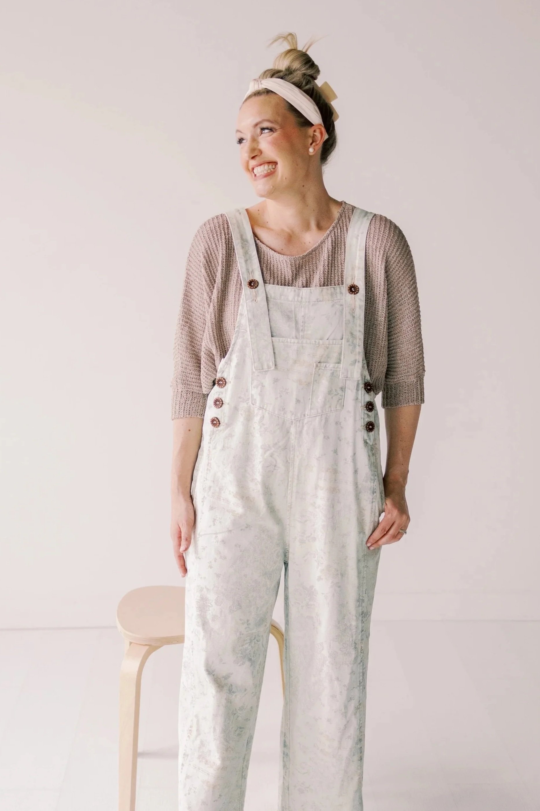 A woman with blonde hair styled in a bun, wearing a white headband and a beige sweater, smiling and looking to the side, wearing light-colored overalls with a floral pattern, and standing beside a small white stool.