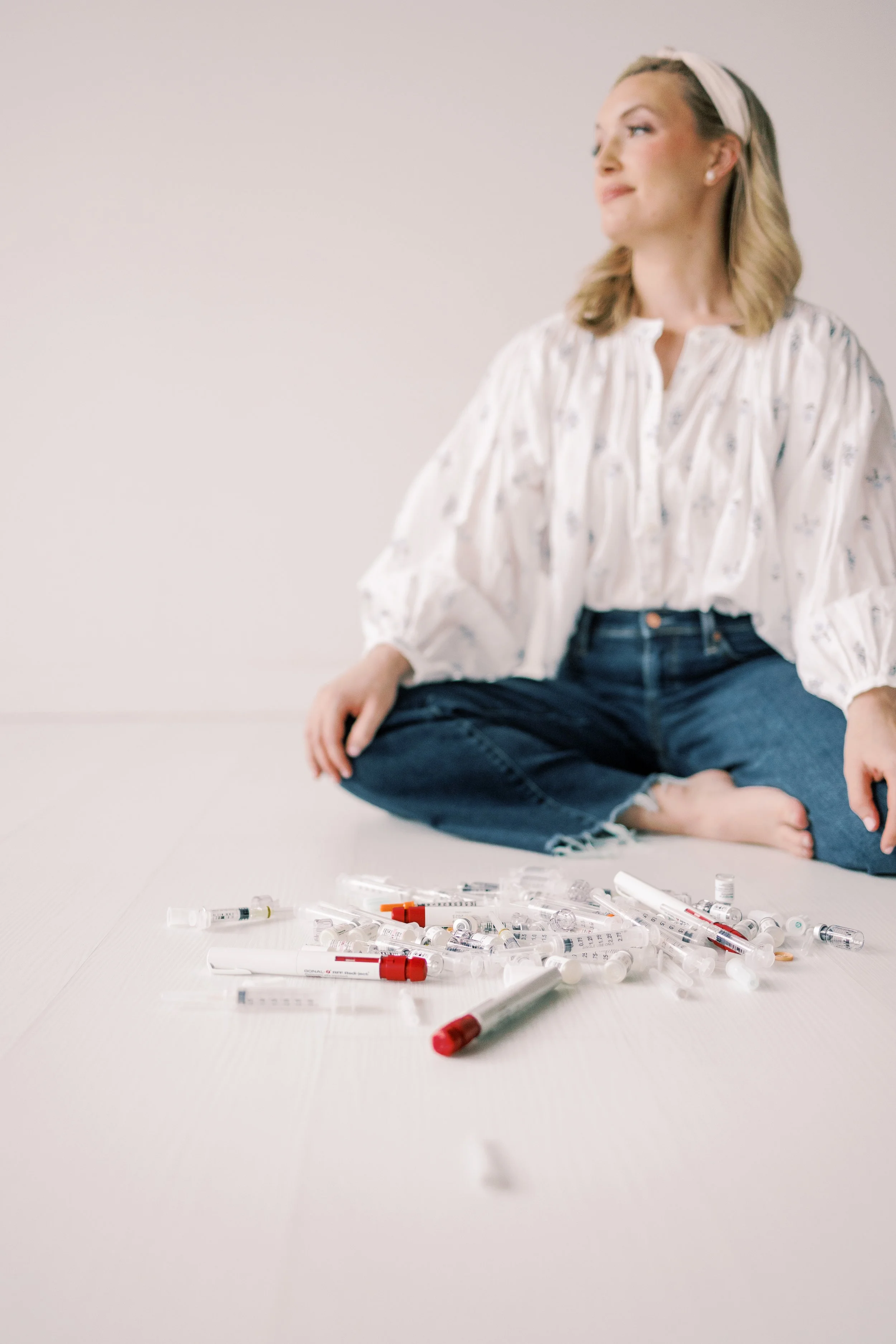 A woman sitting cross-legged on the floor, surrounded by an assortment of medical syringes and vials. She is wearing a white, patterned blouse, blue jeans, a headband, and pearl earrings, with her hair styled in soft waves. The background is plain and light-colored.