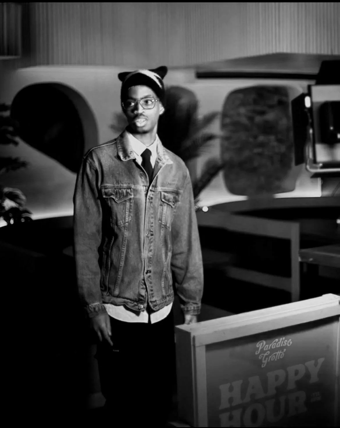 A young man wearing glasses, a beanie with bear ears, a denim jacket, and a shirt with a tie, standing indoors near a sign that says "HAPPY HOUR" and a vending machine, with a painted wall and a potted plant in the background.