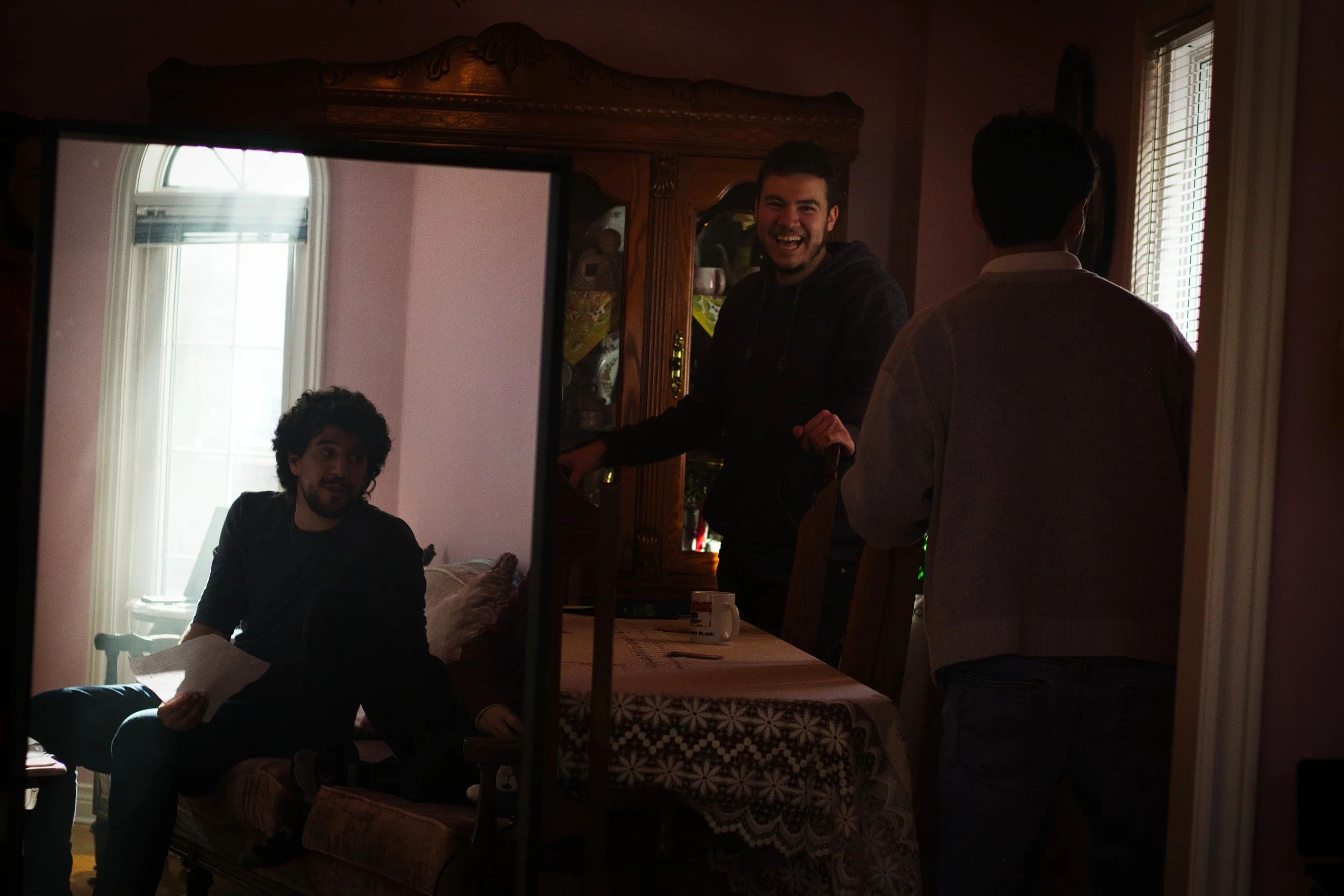 Three men in a dimly lit room with pink walls, one sitting on a sofa with a piece of paper, one standing by a table smiling, and another facing away near a window.