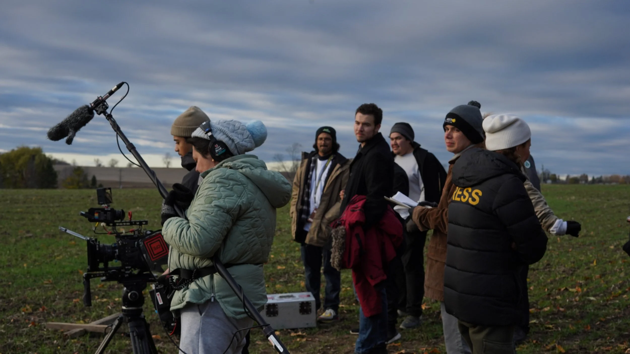 Filmmakers and crew filming outdoors in a field, operating camera and boom microphone, with a group of people standing nearby under a cloudy sky.