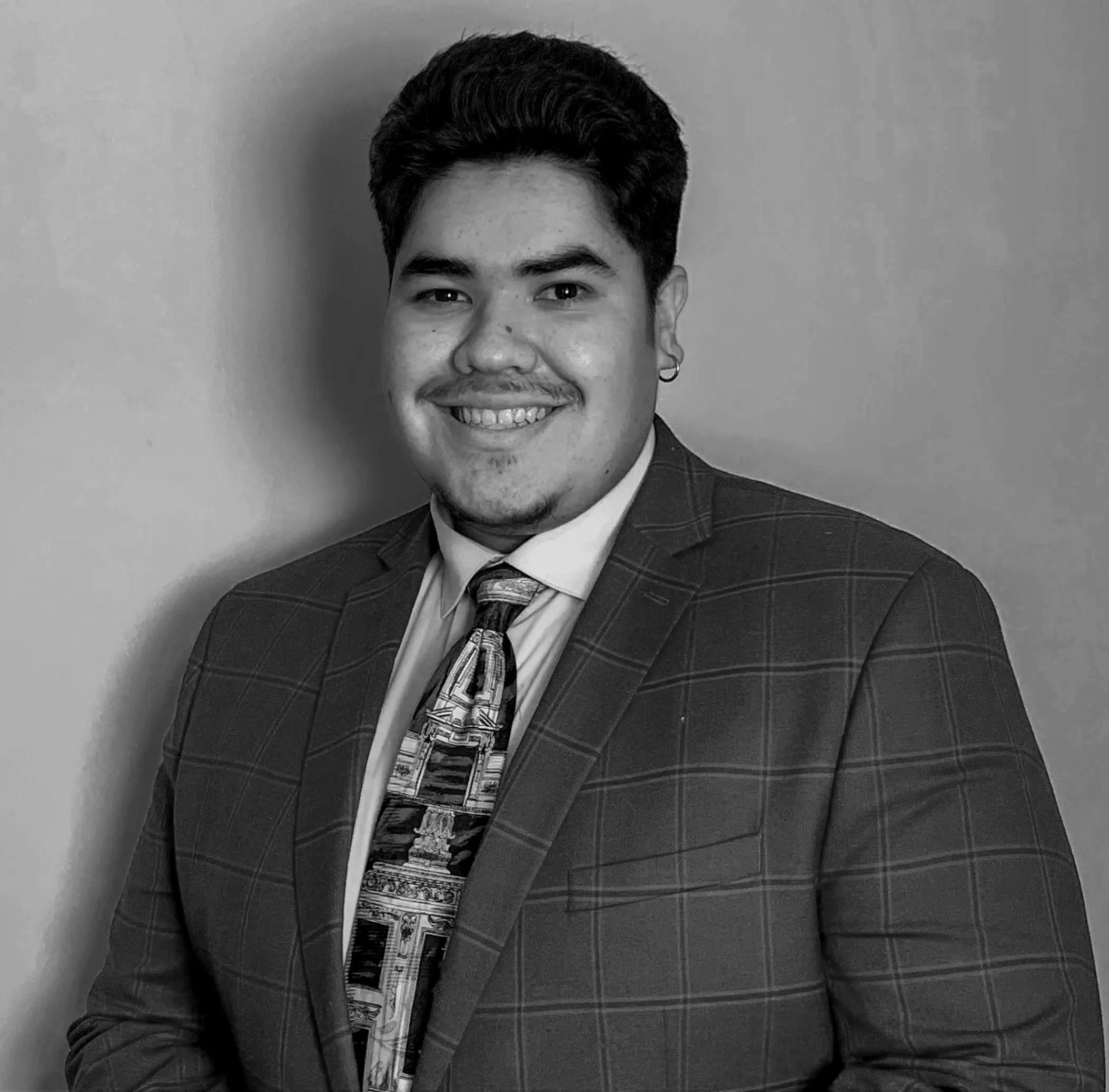 Black and white portrait of a young man smiling, wearing a checkered blazer, dress shirt, and patterned tie, with short dark hair and an earring.