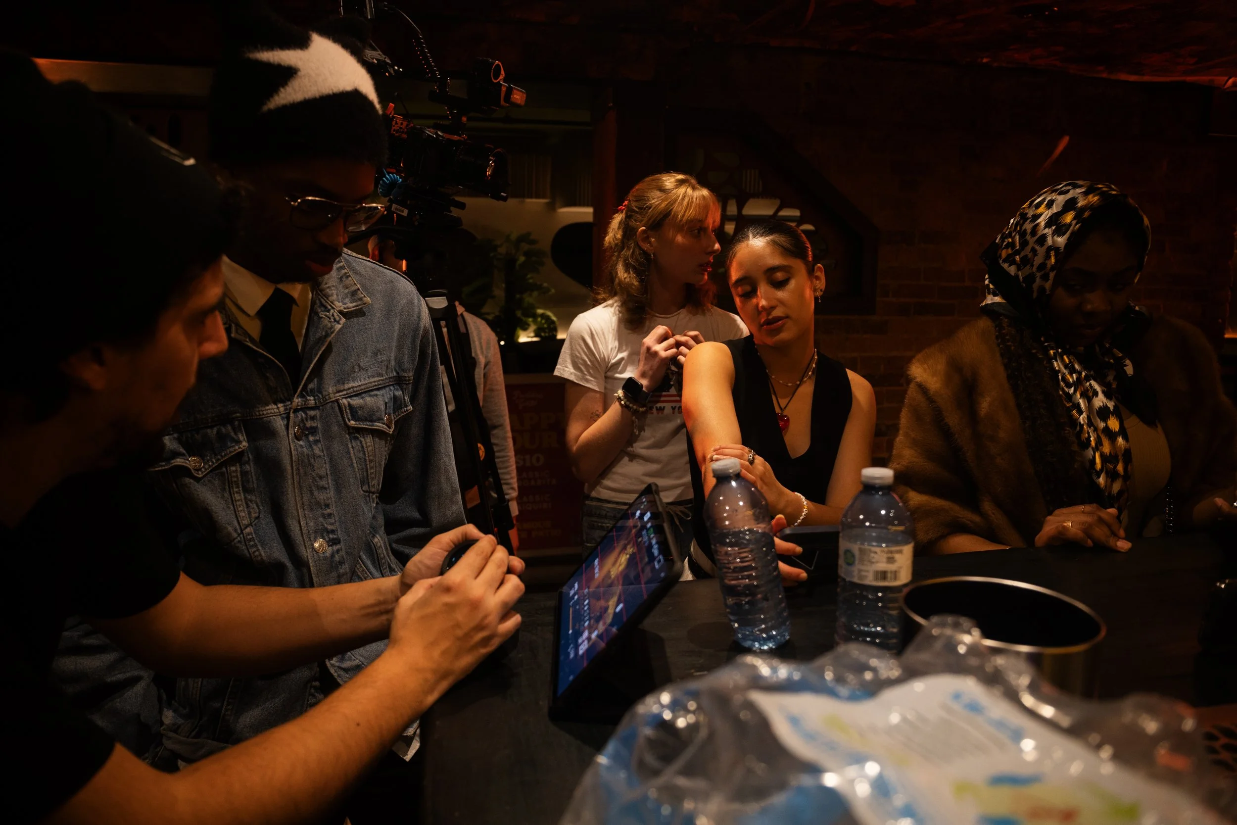 A group of people at a table, some looking at a tablet, in a dimly lit indoor setting.