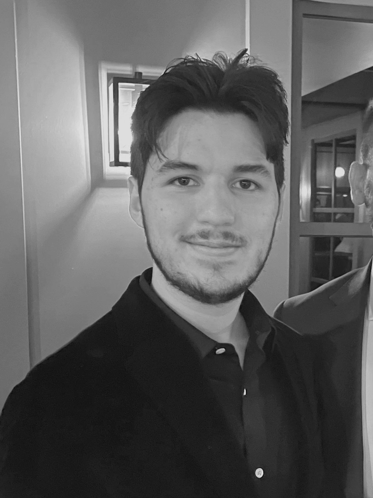 Black and white photo of a young man with dark hair, light facial hair, and a gentle smile, wearing a dark collared shirt, standing indoors with a window and door in the background.