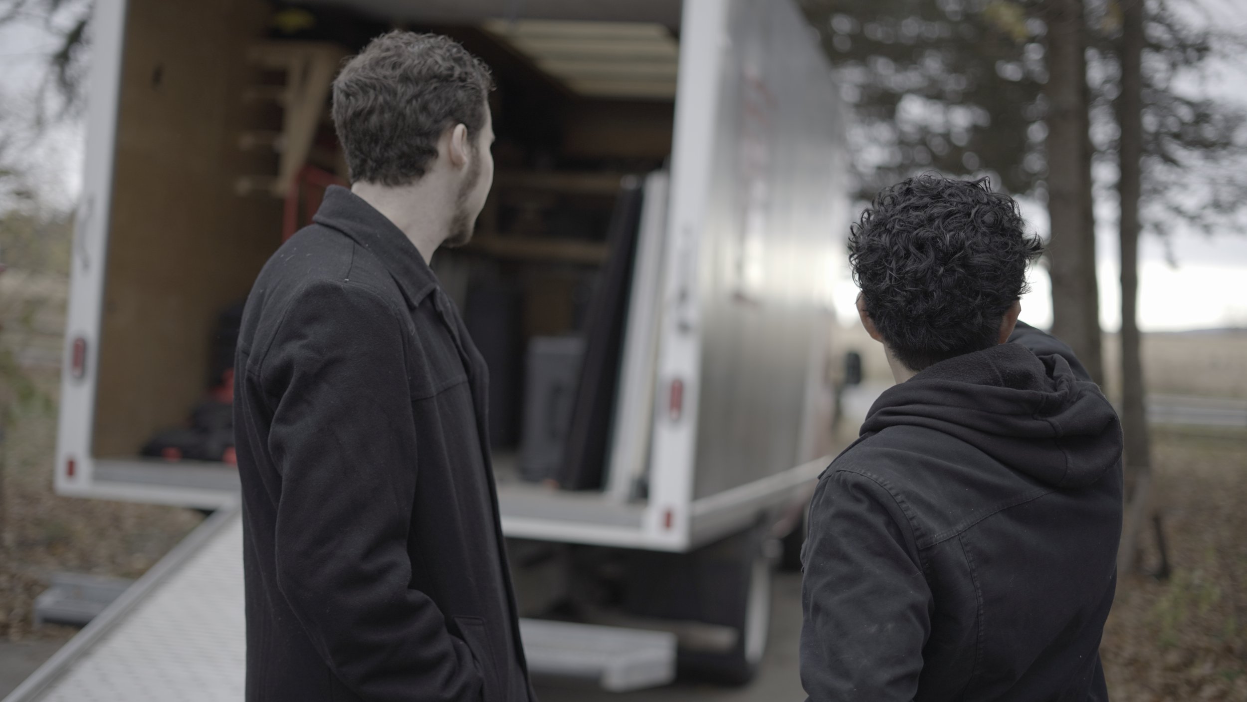 Two men in black jackets standing outdoors near a mobile service or delivery truck, looking at something inside the truck.